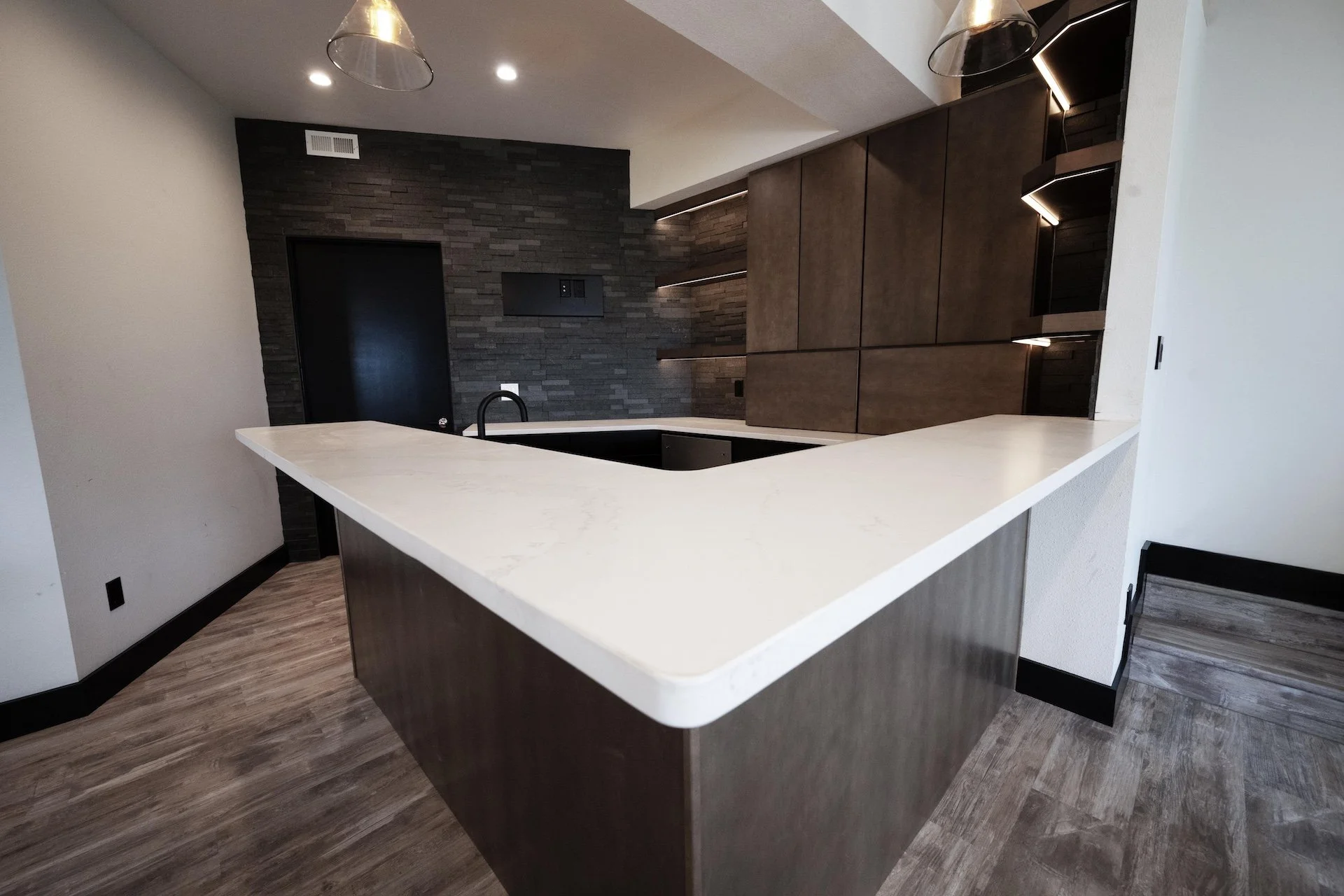 Modern kitchen with white marble countertop, dark wood cabinets, black sink, black faucet, and a textured black accent wall with open shelves and a black door.