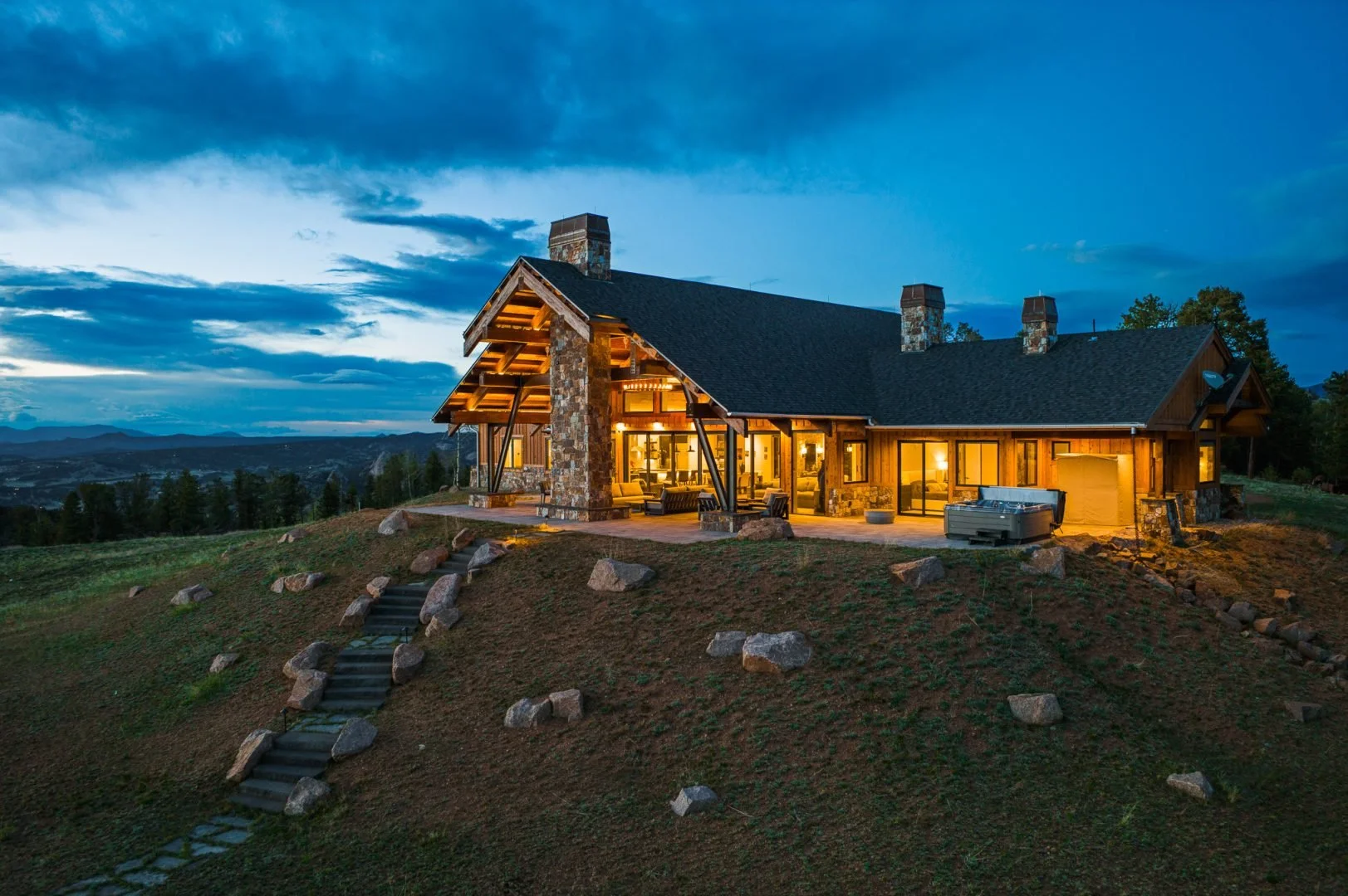 A modern mountain cabin at dusk with large windows, stone and wood exterior, and outdoor lighting, situated on a hill with rocky stairs leading up to it and a scenic landscape in the background.