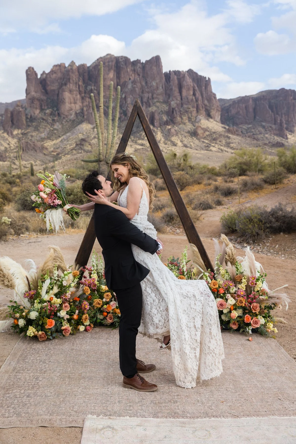 A bride and groom who just got married kissing in front of their lush ceremony triangle shaped arch with an incredible array of ground flowers displayed in front of it in shades that complement the desert such as gold, burnt orange, dusty rose, coral
