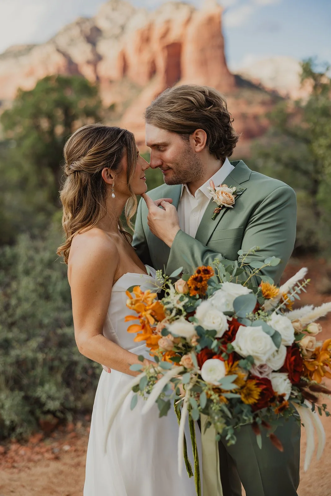 A bride and groom kiss in the Sedona desert.  The groom is wearing a light green suit and the bride is holding a bouquet featuring shades of rust, white, sage and orange.