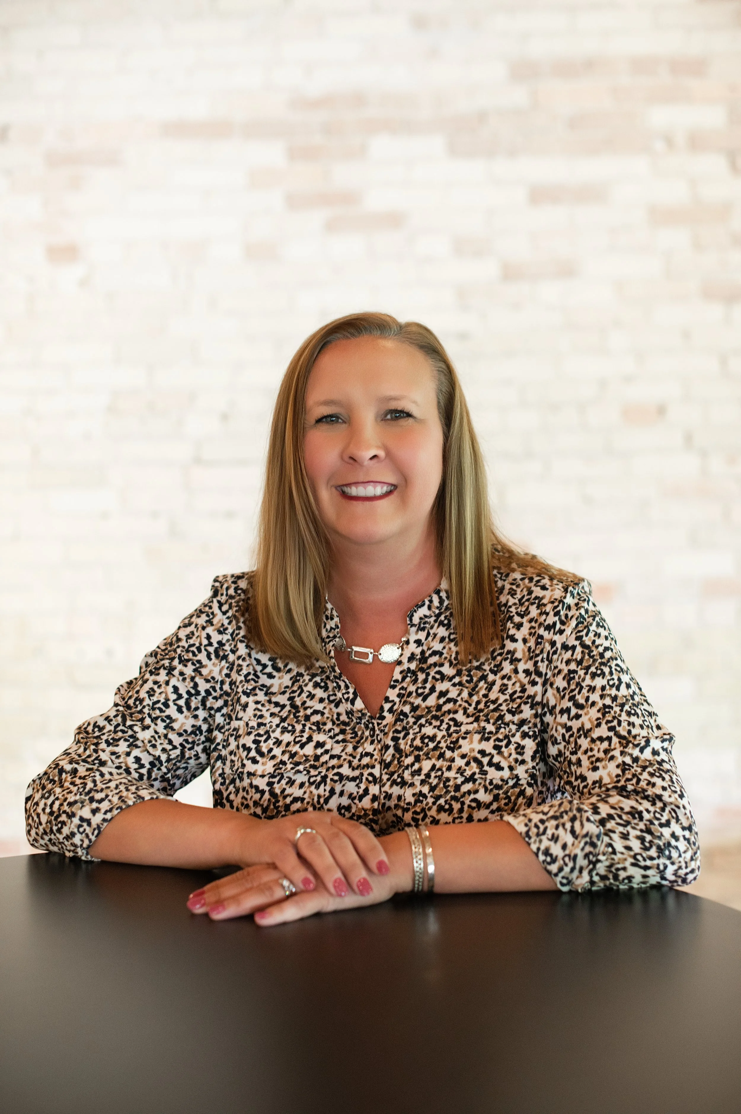 A woman from the Snider & Co real estate team in West Michigan with shoulder-length blonde hair, smiling, sitting at a black table, wearing a leopard print blouse and jewelry, against a white brick wall background.