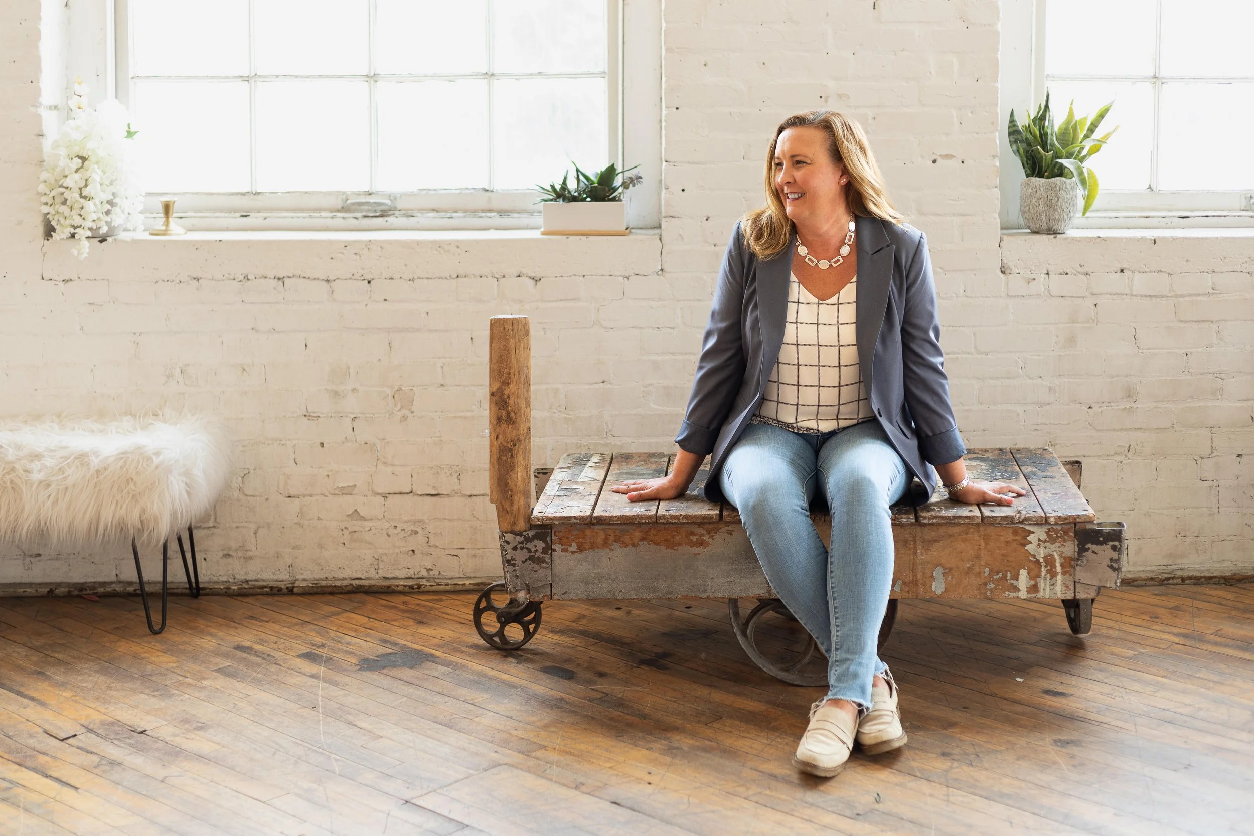 A woman from Snider & Co estate services in West Michigan sitting on a rustic wooden cart in a room with white brick walls and wooden floors, smiling and looking to her left, with potted plants on the windowsill behind her.