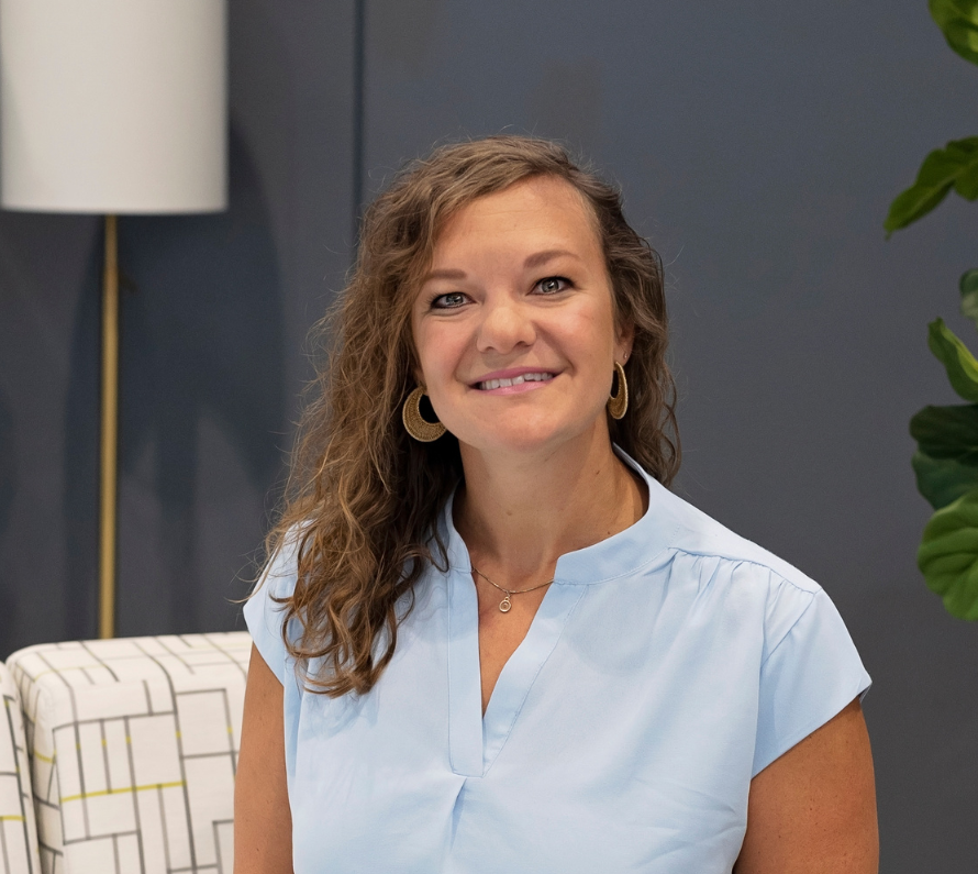 A woman with curly brown hair smiling, wearing a light blue blouse, gold earrings, and a necklace, sitting in a modern office space with a patterned chair, a white floor lamp, and green plants in the background.