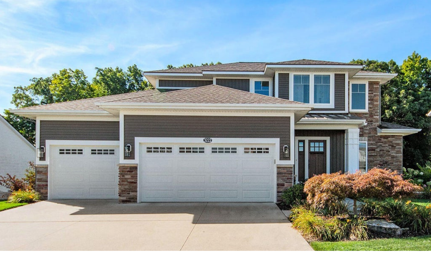 Front view of a modern two-story house with a three-car garage, gray siding, white trim, stone accents, and landscaped garden with bushes and trees. It is a home Snider & Co. real estate services sold.