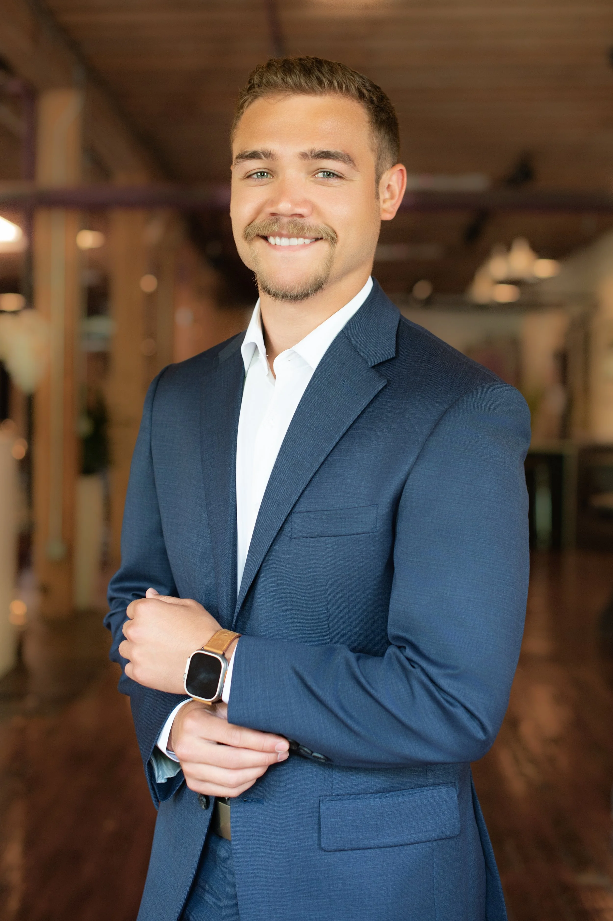 A young man with short brown hair, a light beard, and blue eyes, smiling and dressed in a navy blue suit with a white shirt, standing in a warmly lit indoor space.