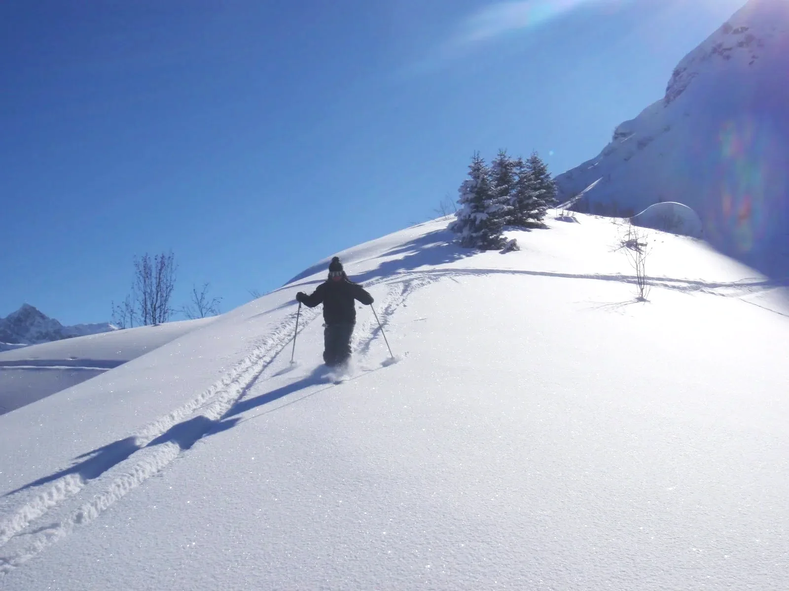 Une personne en vêtements d'hiver fait de la raquette dans la neige sur une colline en montagne, avec des arbres enneigés et un ciel bleu clair.