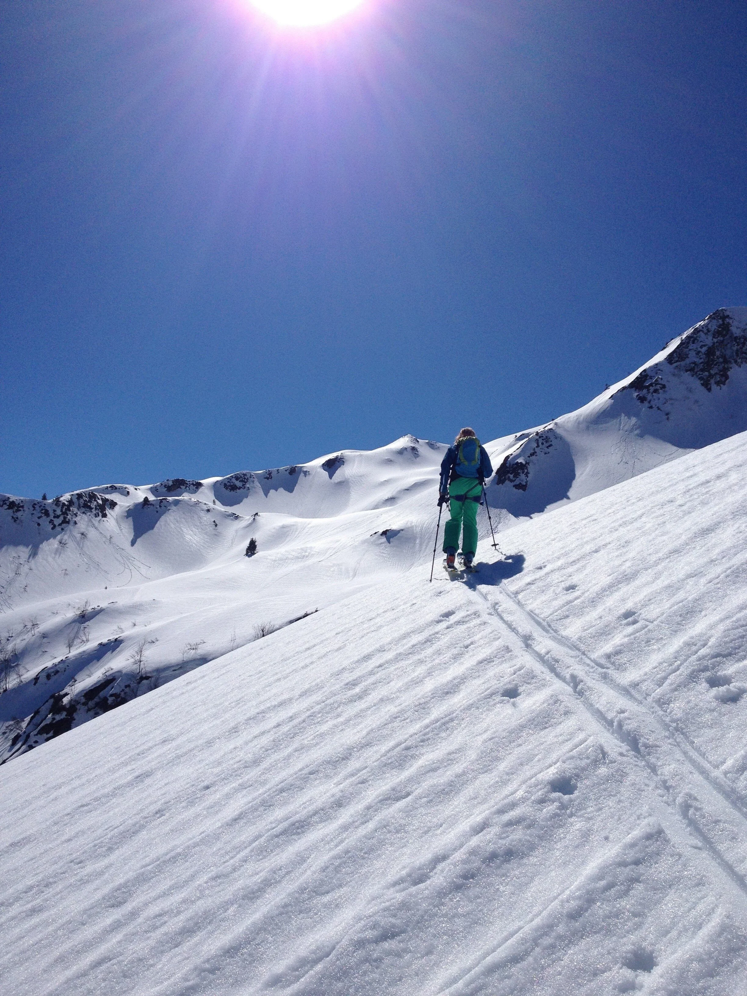 Une personne en vêtements de ski marche dans la neige en montagne sous un ciel bleu et ensoleillé.
