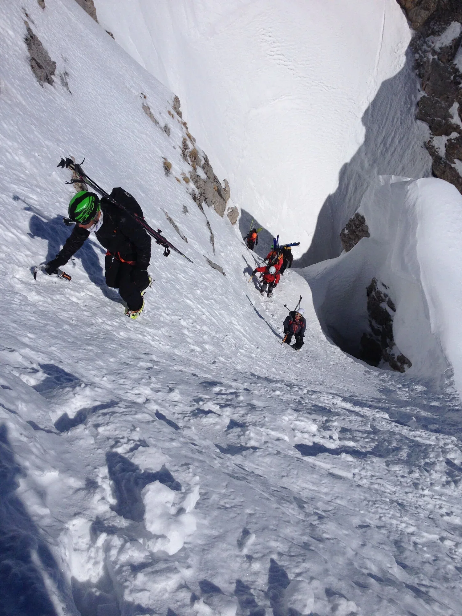 Groupe d'alpinistes escaladant une pente enneigée dans une gorge de montagne, avec des falaises de roche autour.