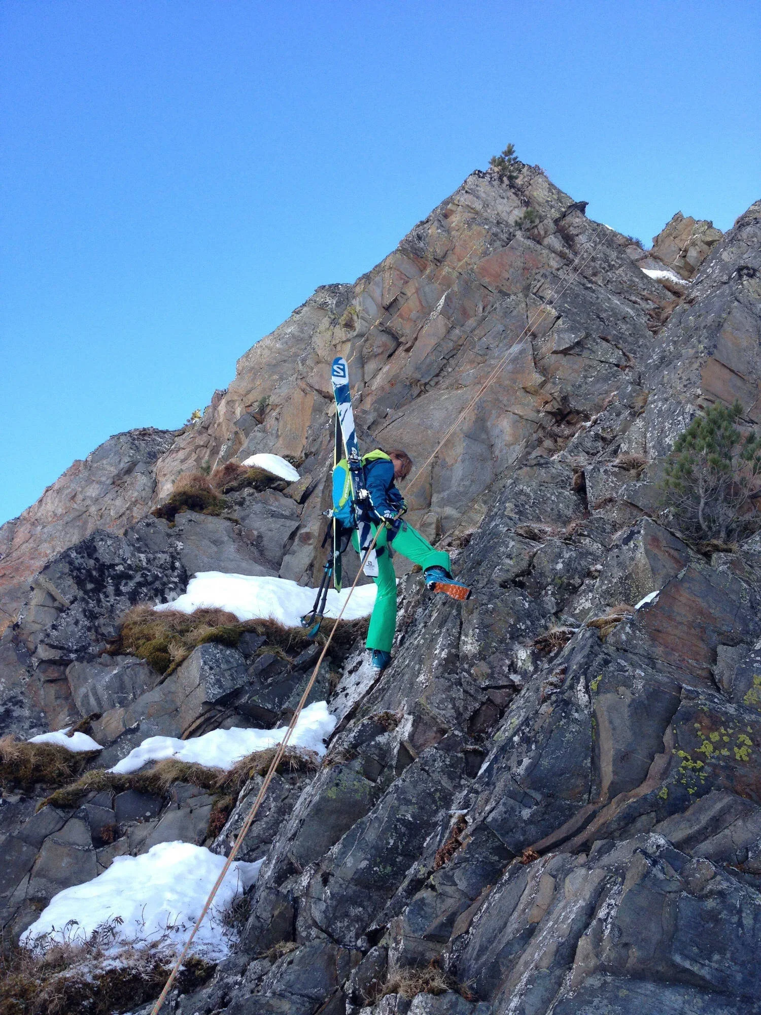 Une personne fait de l'alpinisme sur une face rocheuse escarpée avec de la neige, portant un sac à dos et des skis attachés à ses épaules, dans un environnement montagneux.