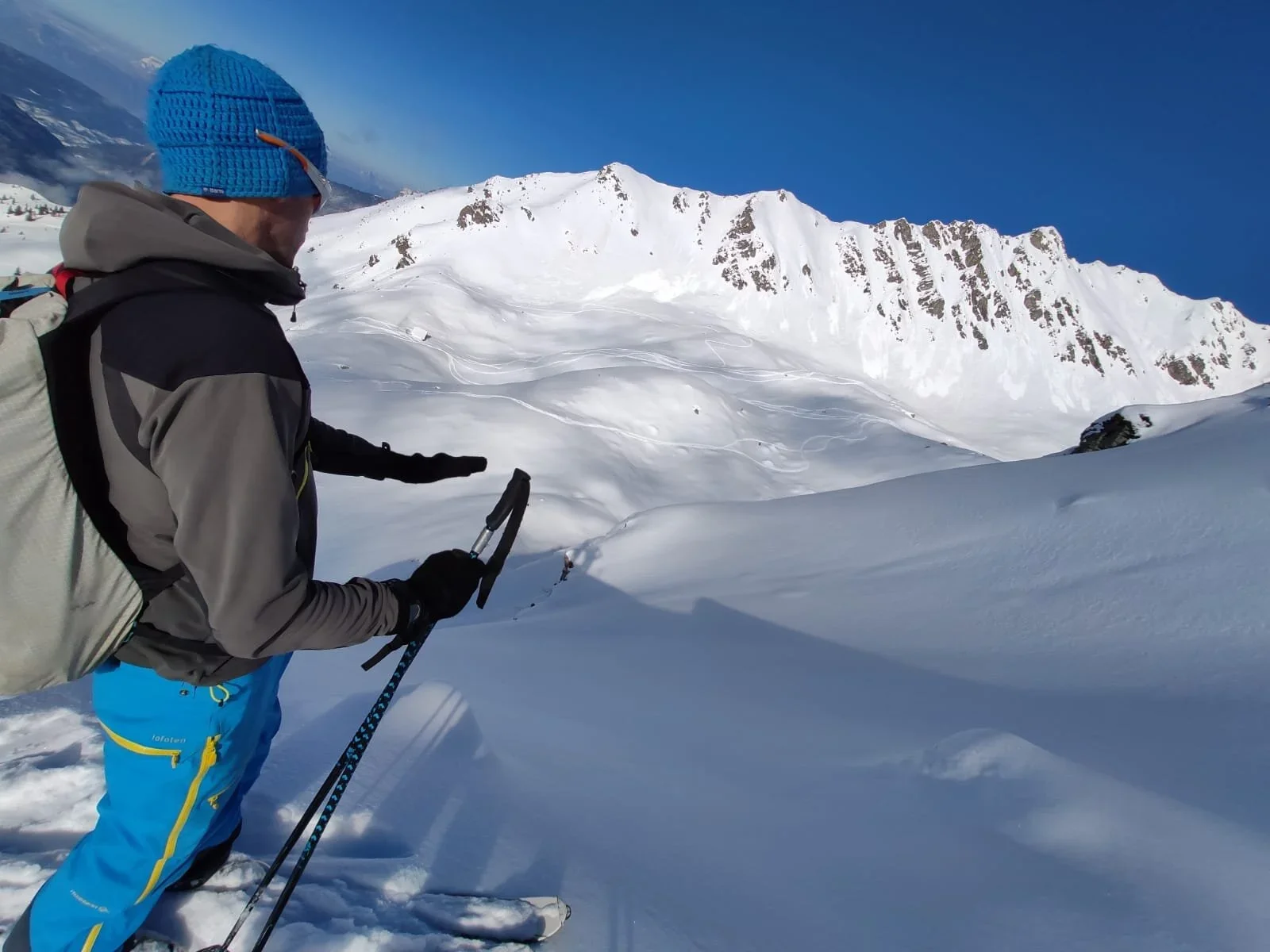 Skieur en montagne enneigée portant un casque bleu et des vêtements colorés, avec des skis et des bâtons, face à un paysage de montagnes enneigées sous un ciel bleu.