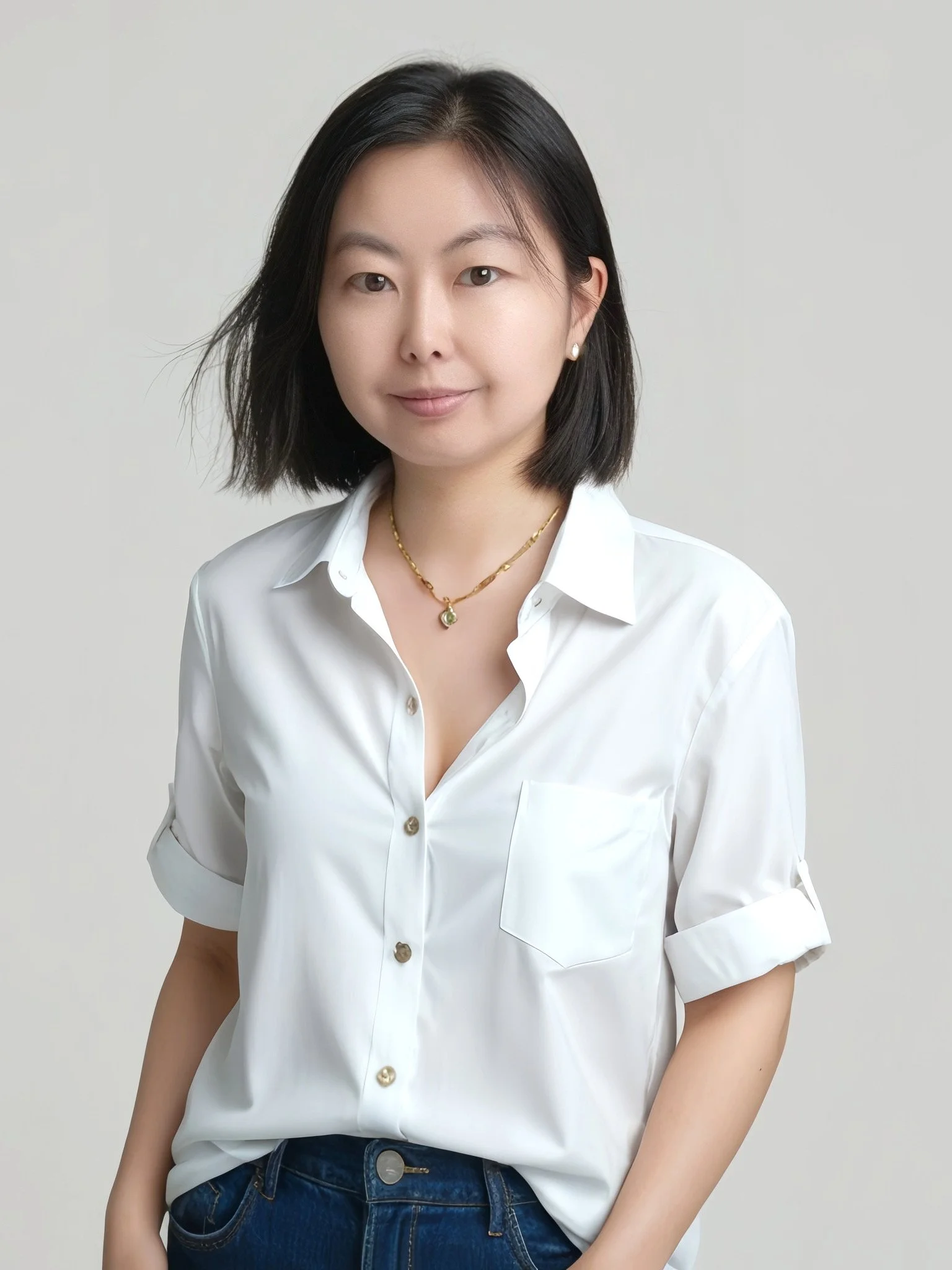 A woman with shoulder-length black hair wearing a white button-up shirt with rolled-up sleeves, blue jeans, a gold necklace, and pearl earrings, standing against a plain neutral background.