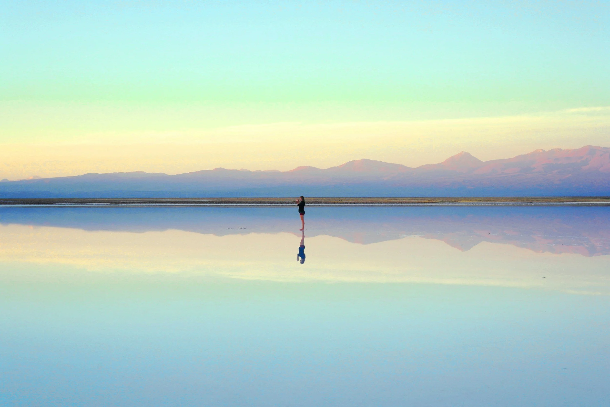 A person standing on a reflective salt flat, with mountains and a pastel-colored sky in the background, creating a mirror-like effect.