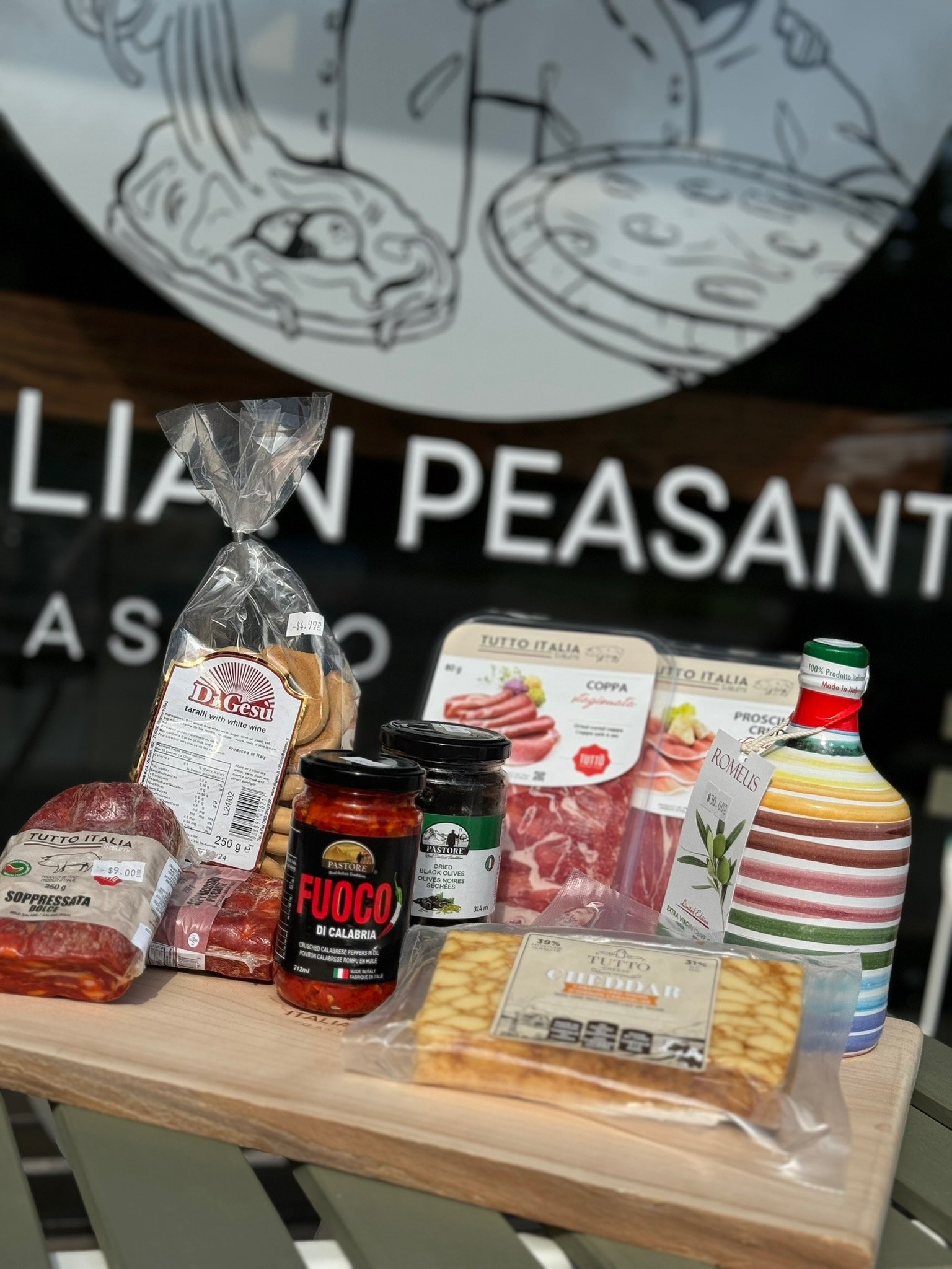 Various Italian food products, including pasta, canned olives, canned tomatoes, meat, and jarred sauces, arranged on a wooden table outside, with a sign reading 'Italian Peasant' in the background.