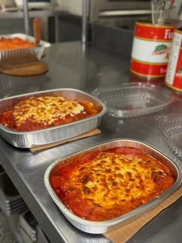 Two aluminum containers of baked lasagna with melted cheese on top on a kitchen counter. In the background, there are canned tomato products and a clear plastic lid.