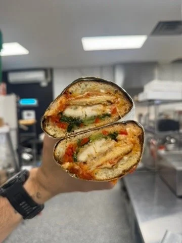 Hand holding a halved sandwich with visible layers of chicken, cheese, tomato, and green leafy vegetables, in a commercial kitchen setting.