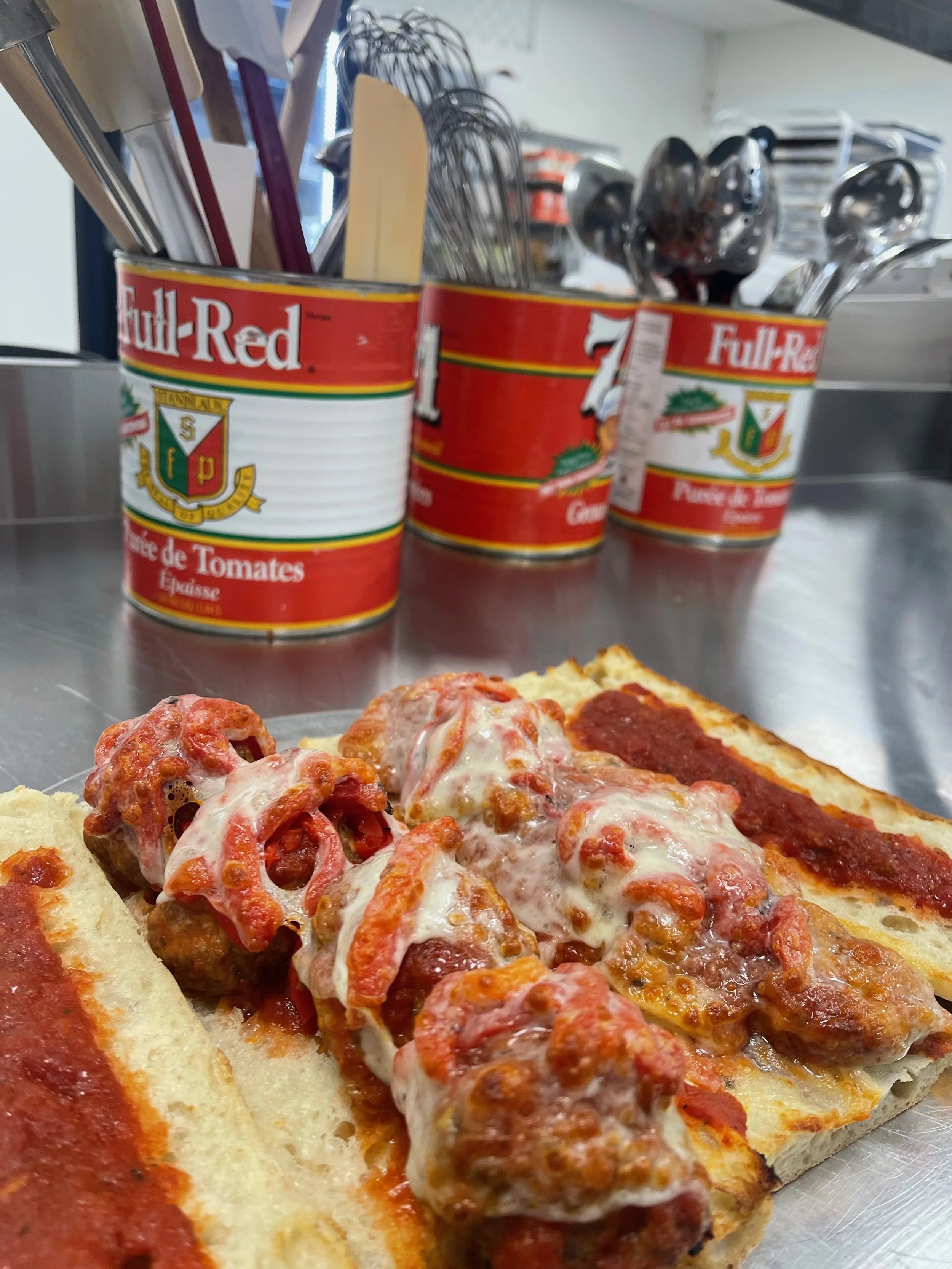Slices of pizza with meatballs and marinara sauce on a stainless steel countertop, with three cans of tomato paste and can lids in the background.