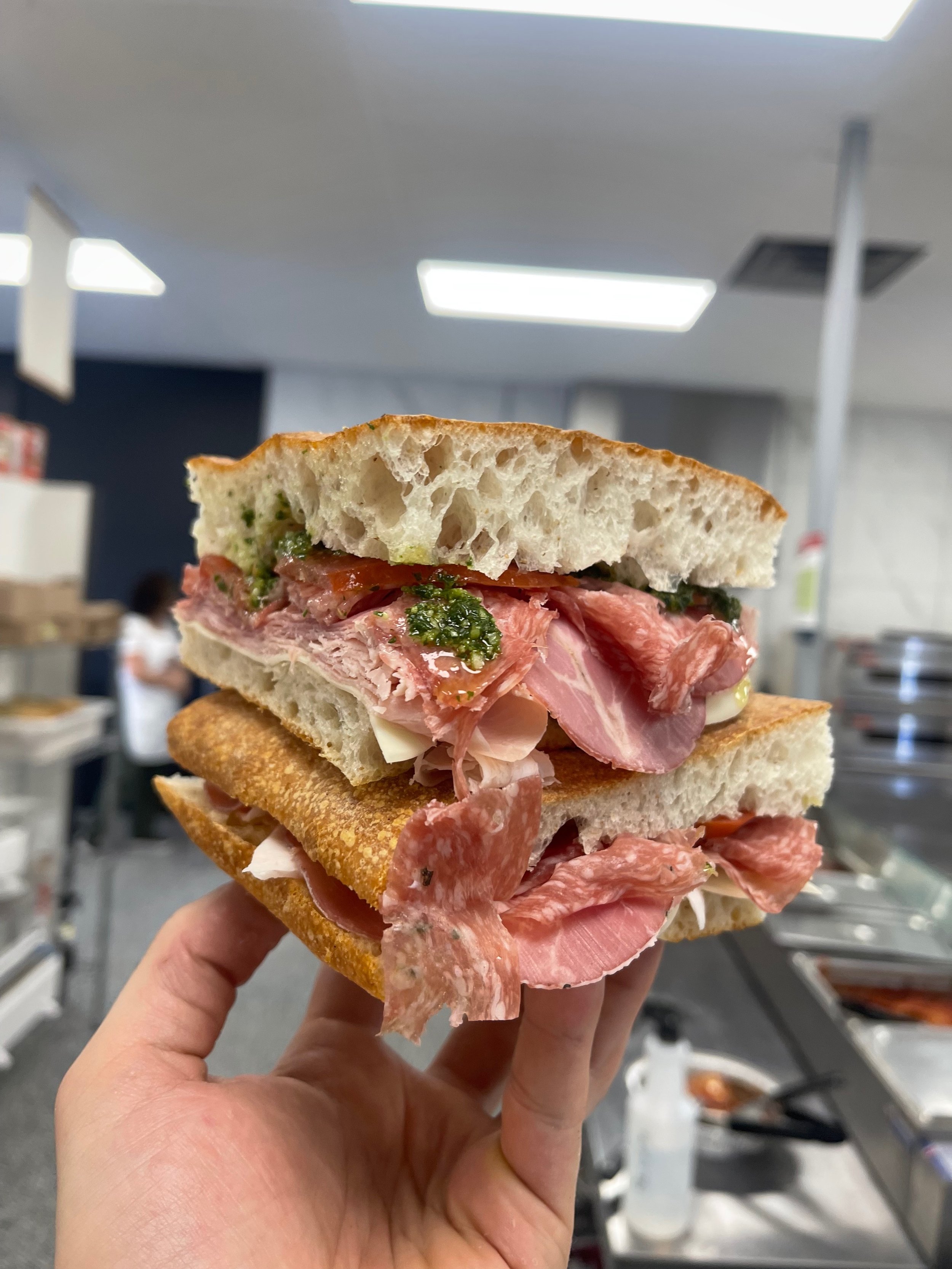 Hand holding a large sandwich with various meats and greens inside, in a cafeteria setting.