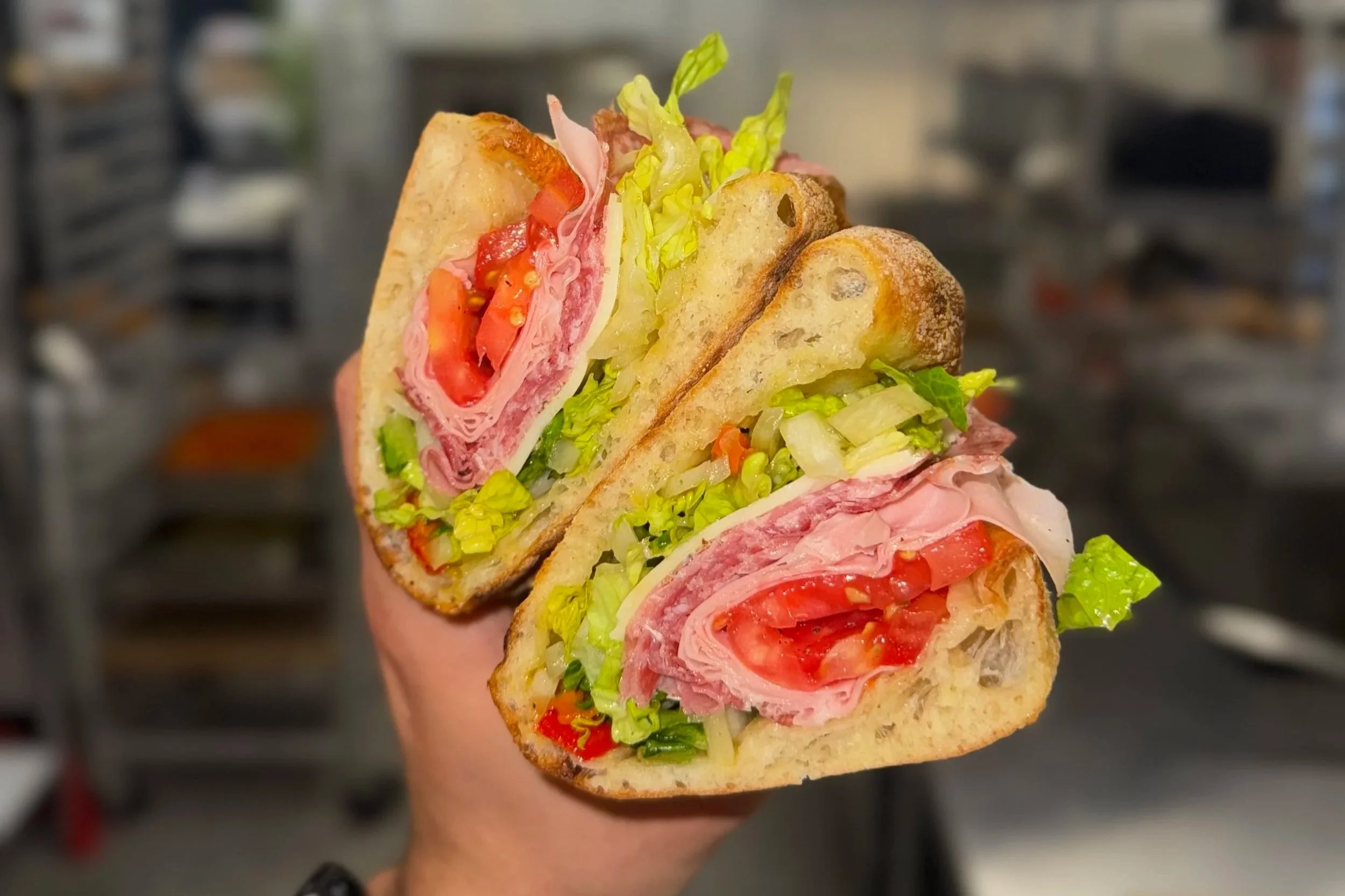 Close-up of a hand holding a sandwich with turkey, tomatoes, lettuce, and toasted bread, with a blurred kitchen background.