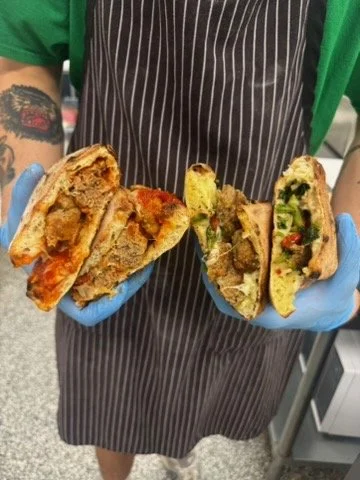 Person holding two halves of a sandwich with melted cheese, meat, lettuce, tomato, and other vegetables, wearing a striped apron and blue gloves.