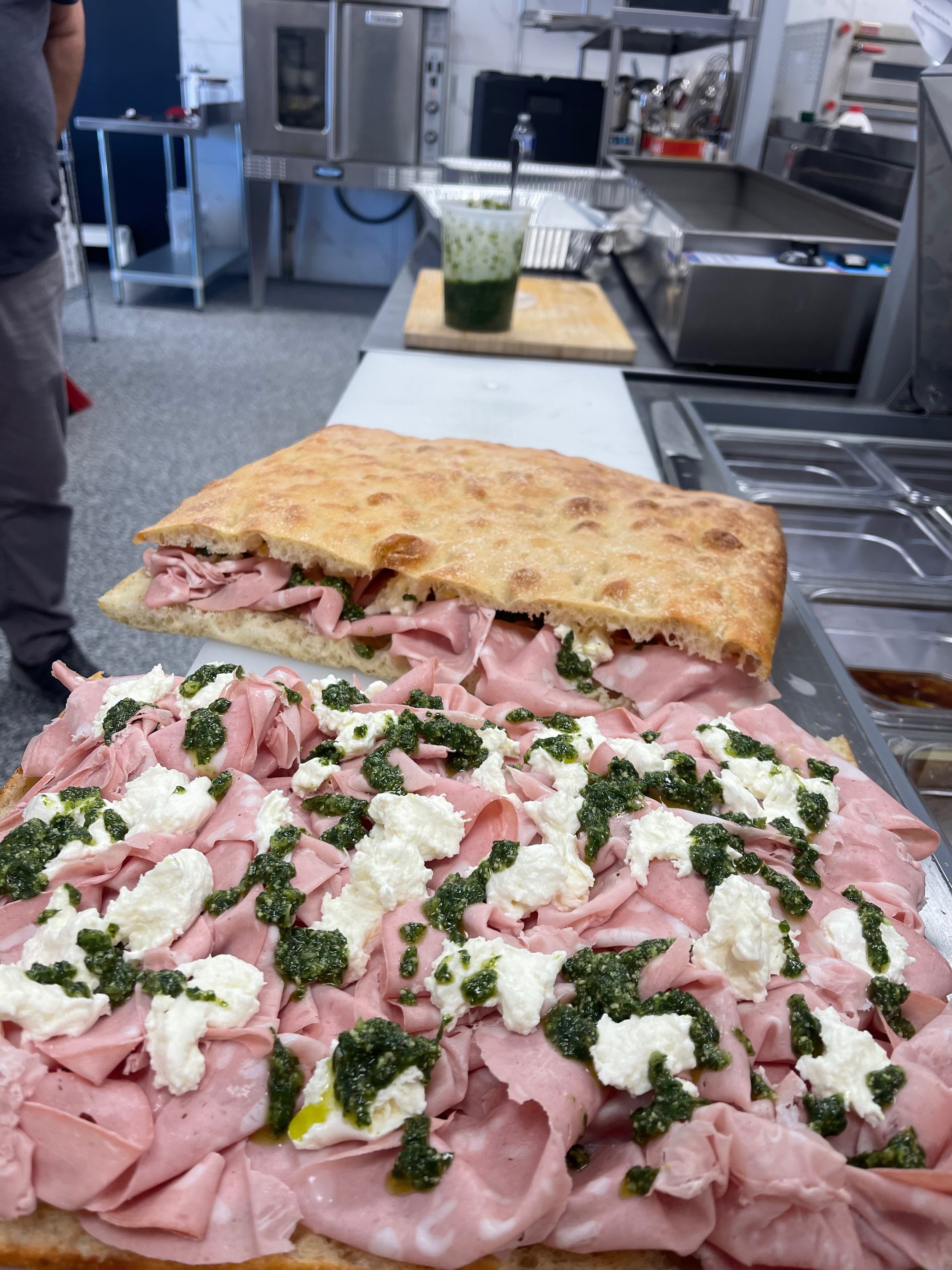 A large sandwich with ham, mozzarella cheese, and green pesto on focaccia bread, served on a prep table in a commercial kitchen.