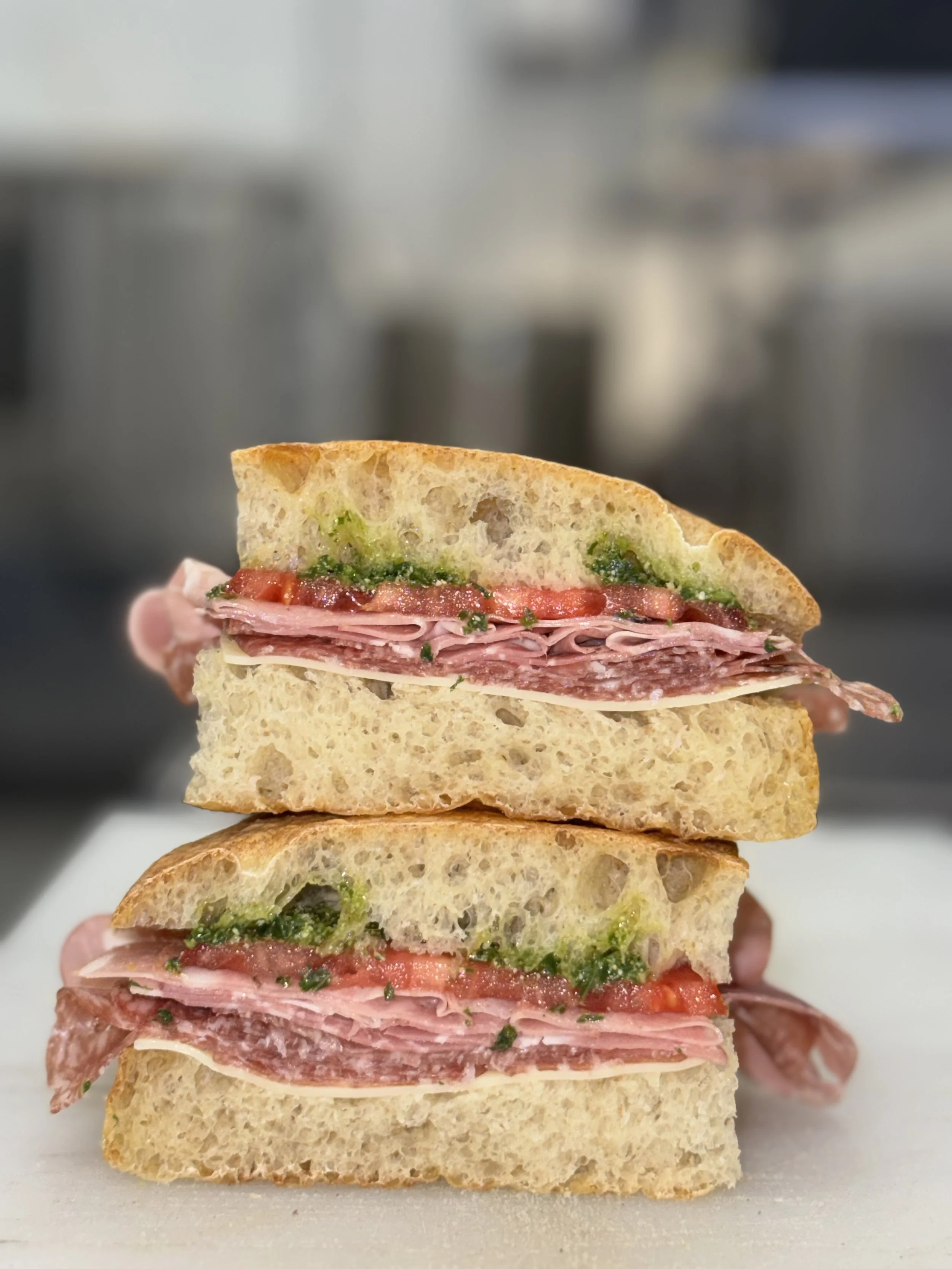 Stacked ham and salami sandwich with tomato, green sauce, and bread in a kitchen setting.