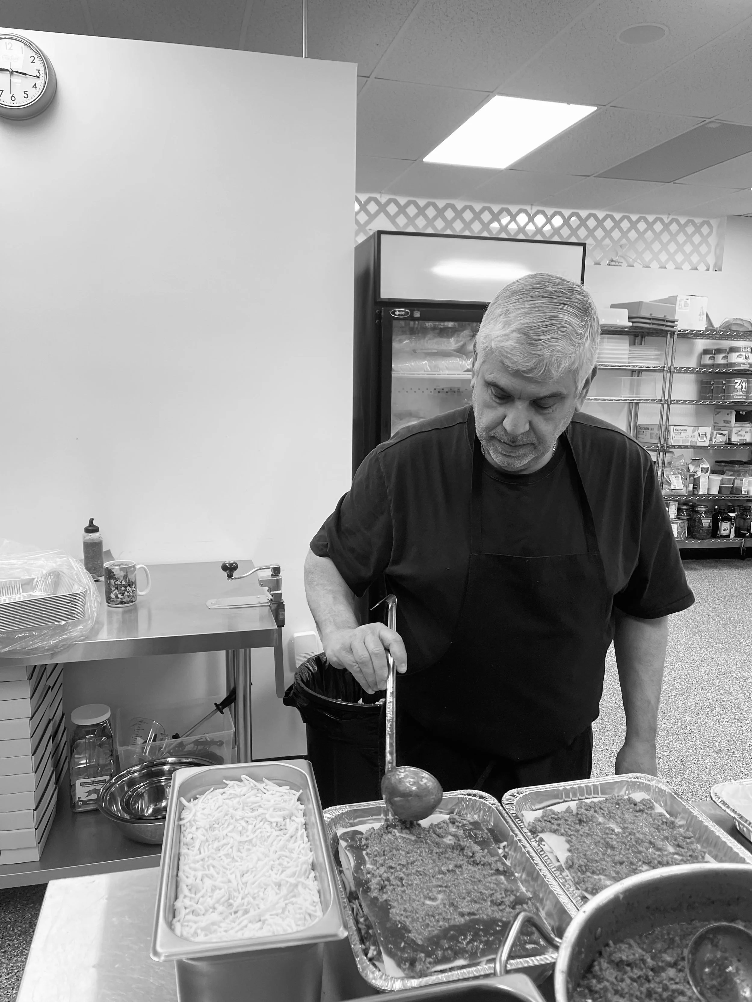 A man cooking food with a ladle in a kitchen or food service setting, with trays of prepared food and various kitchen tools around him.