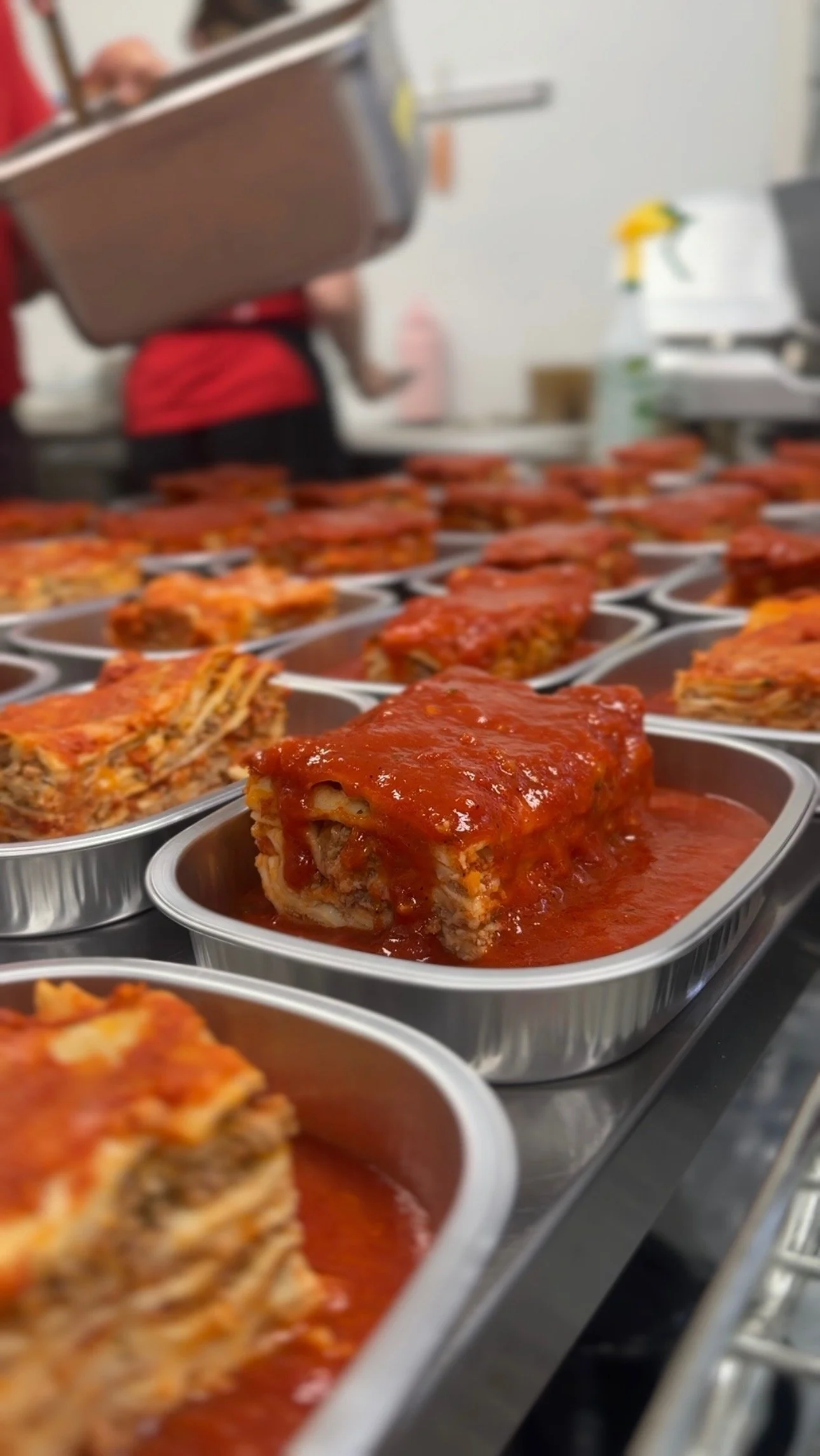 Tray of prepared lasagna with tomato sauce on a kitchen counter, with a person in the background holding a tray and other kitchen items.