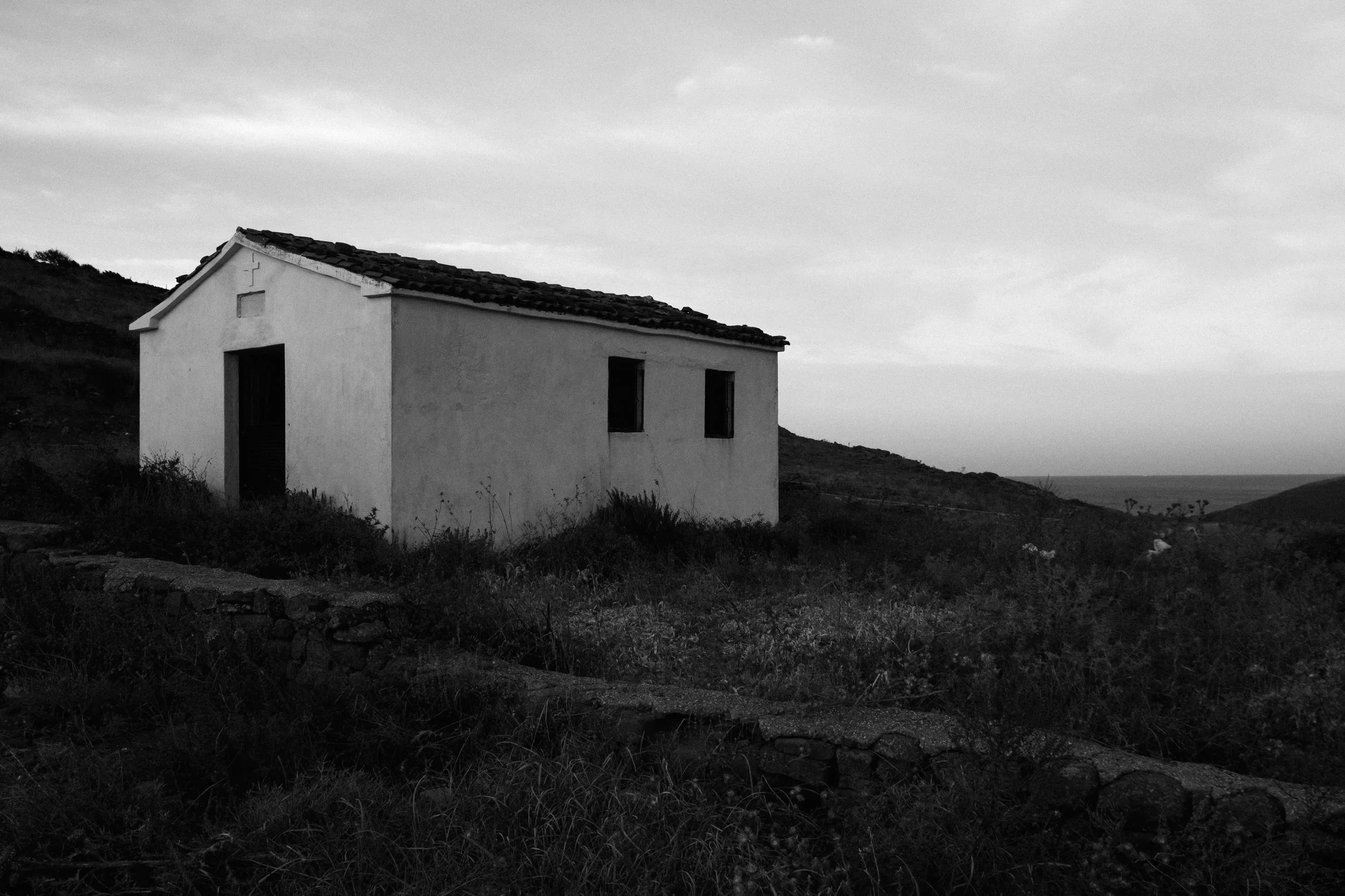 A small white house with a tiled roof on a hillside, surrounded by low vegetation, in black and white.