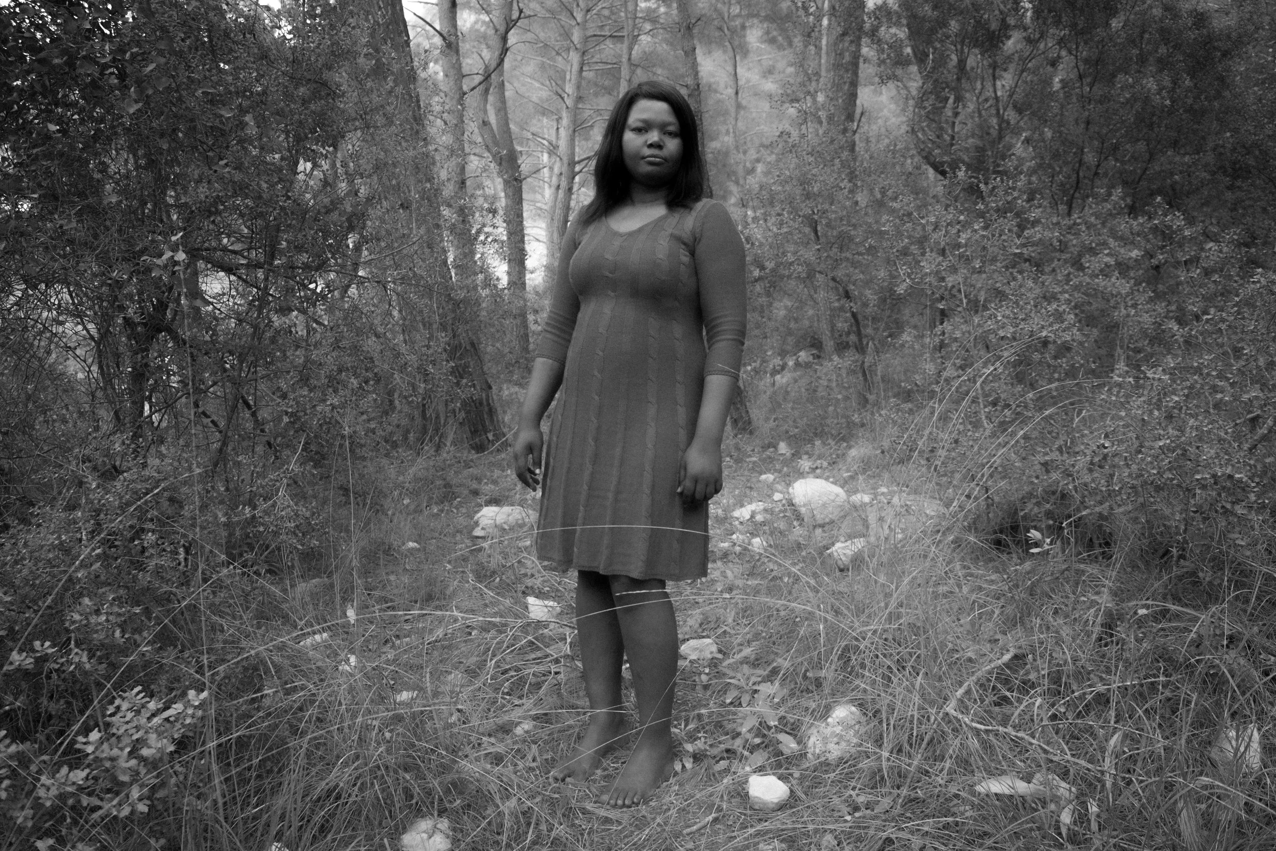 A woman standing barefoot in a wooded outdoor area, wearing a knee-length dress with long sleeves, surrounded by grass and trees.