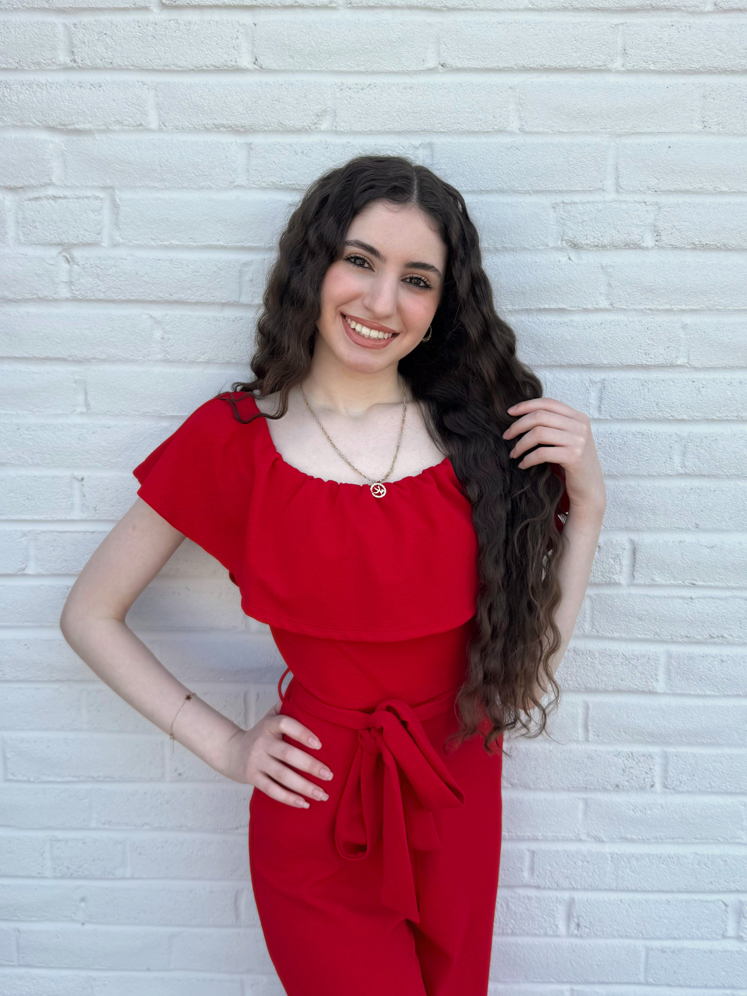 A young woman with long curly hair smiling and standing against a white brick wall, wearing a pink top with pearl embellishments and blue jeans.