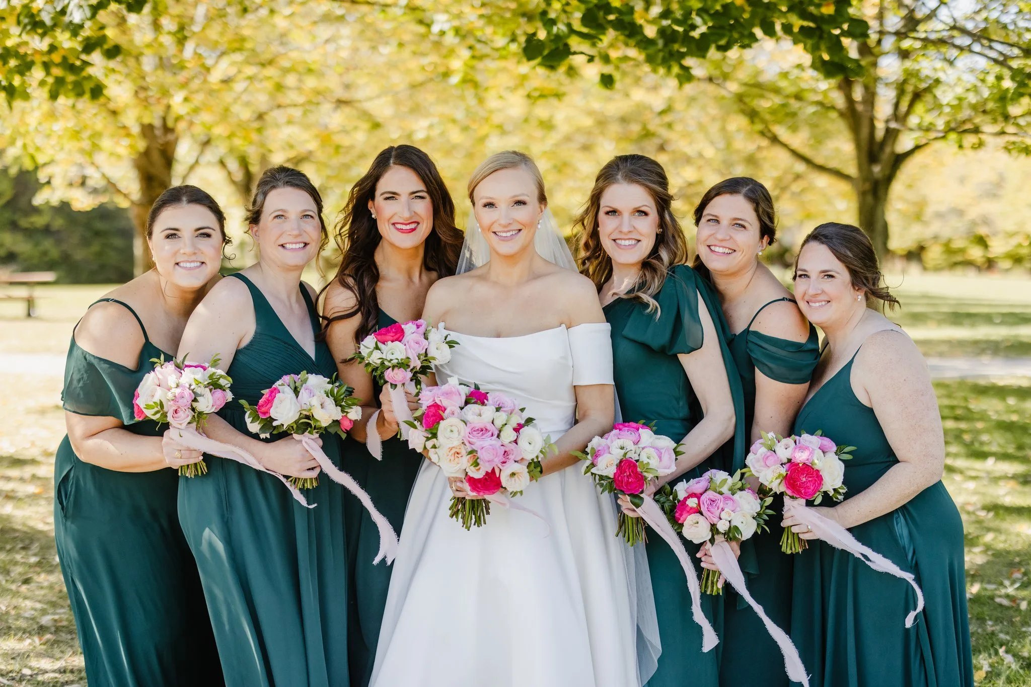 A group of seven women, one in a wedding dress and the others in matching green dresses, standing outdoors under trees with autumn leaves, holding bouquets of pink and white flowers.