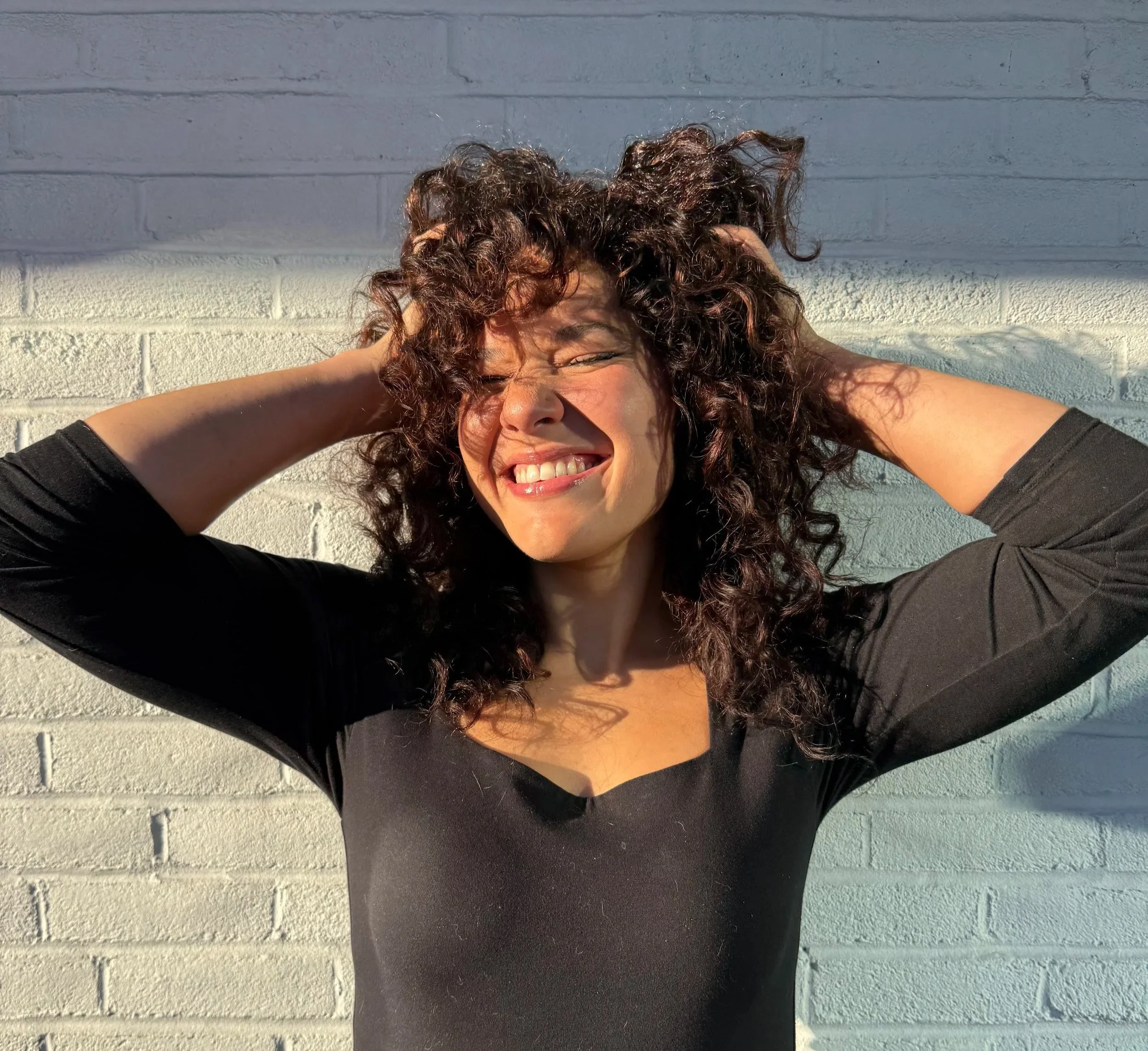 A woman with curly hair smiling with her eyes closed, leaning against a white brick wall with her arms behind her head, wearing a black top.