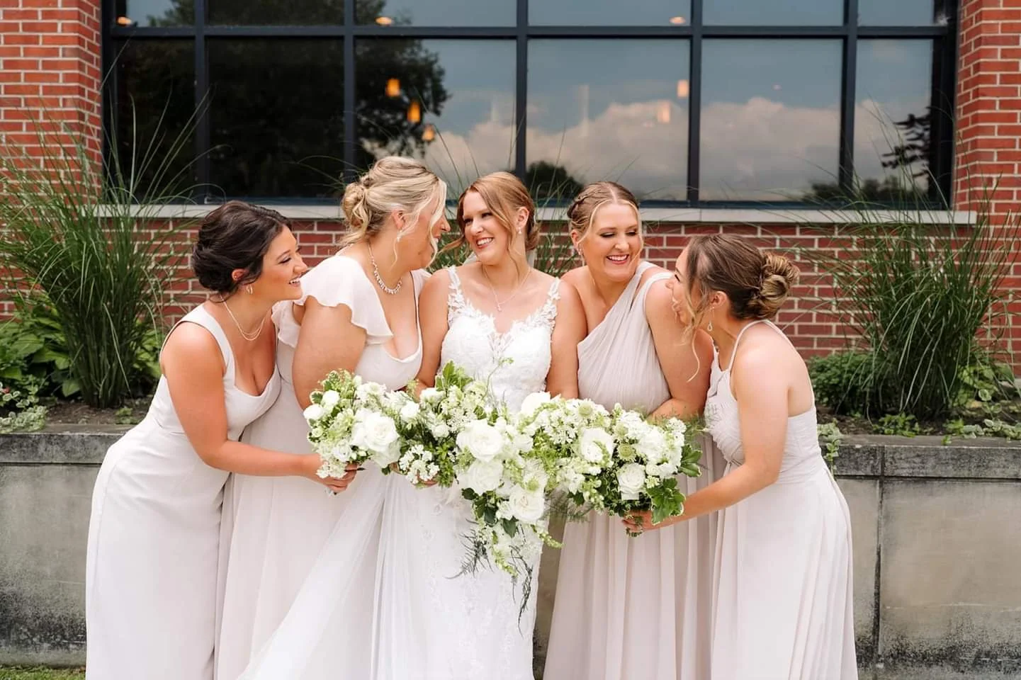 A bride and five bridesmaids smiling and holding bouquets of white flowers outside in front of a brick building.