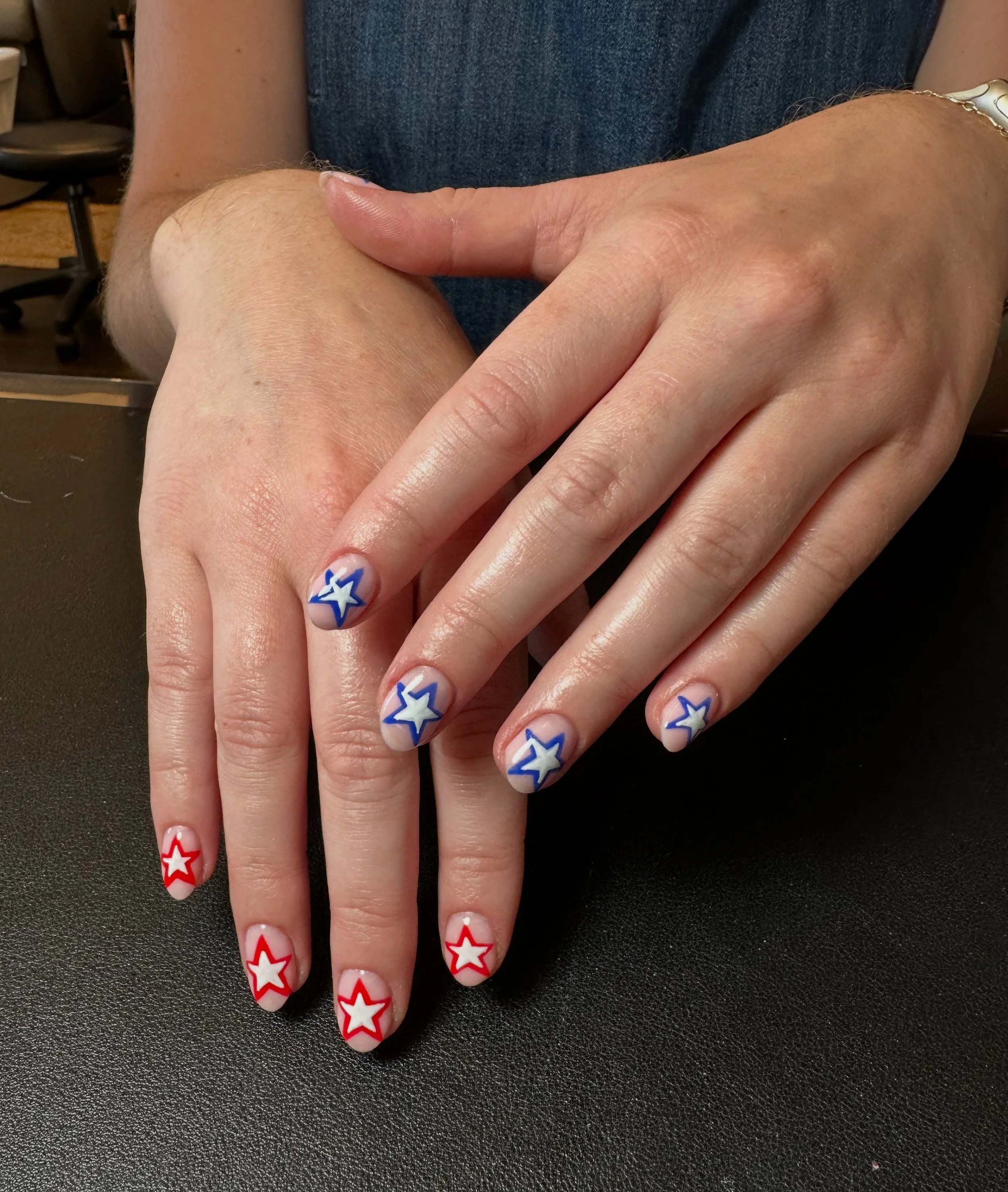 Close-up of two hands with star-patterned red, white, and blue nail art, resting on a black surface, with a person wearing a dark blue shirt in the background.