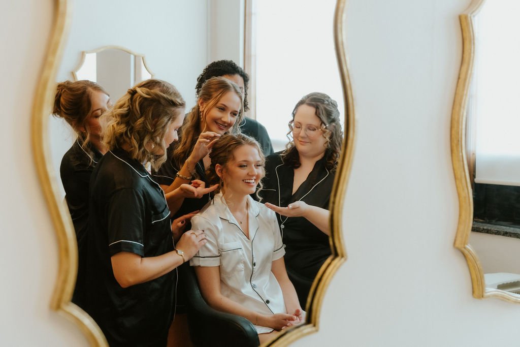 A woman having her hair done by hairstylists, while being reflected in a decorative mirror, in a room with natural light.