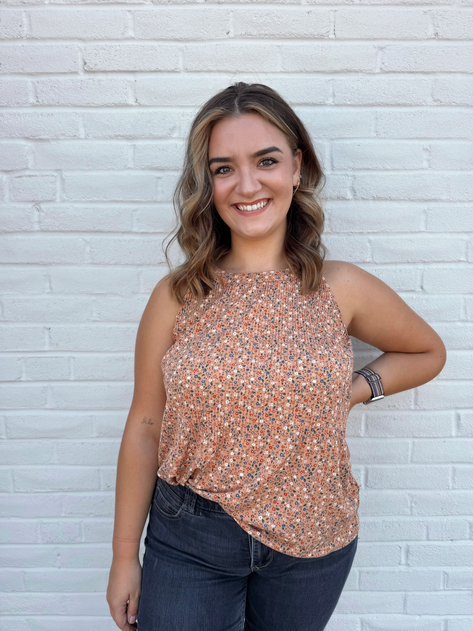 A young woman with blonde hair and a big smile, wearing a brown blouse with ruffled sleeves and high-waisted blue jeans, standing against a white brick wall.
