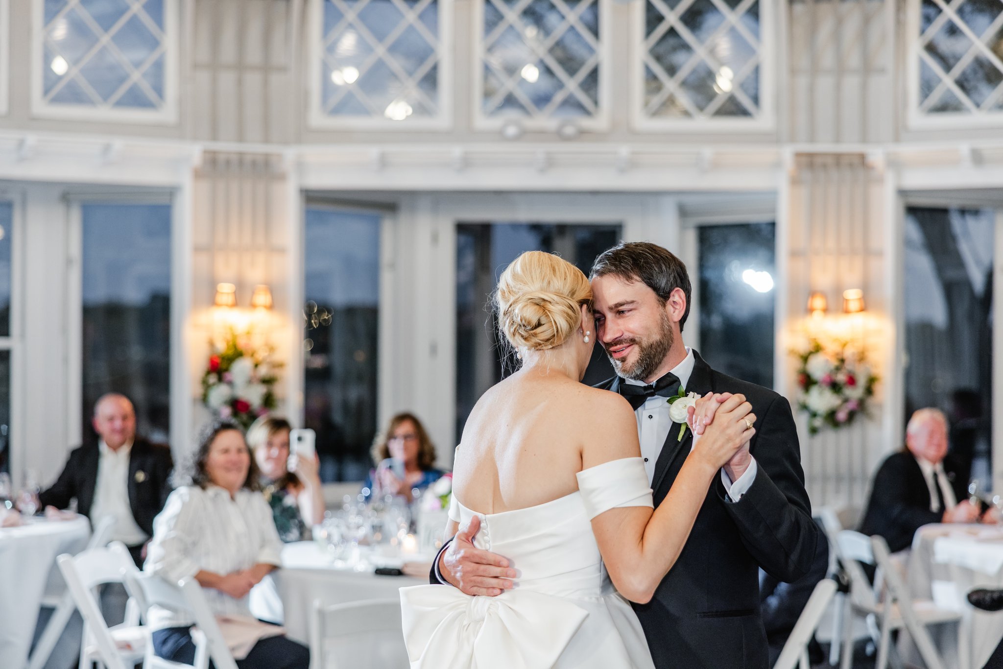 A bride and groom share a dance at their wedding reception, surrounded by seated guests in an elegant, well-lit venue with large windows and floral decorations.