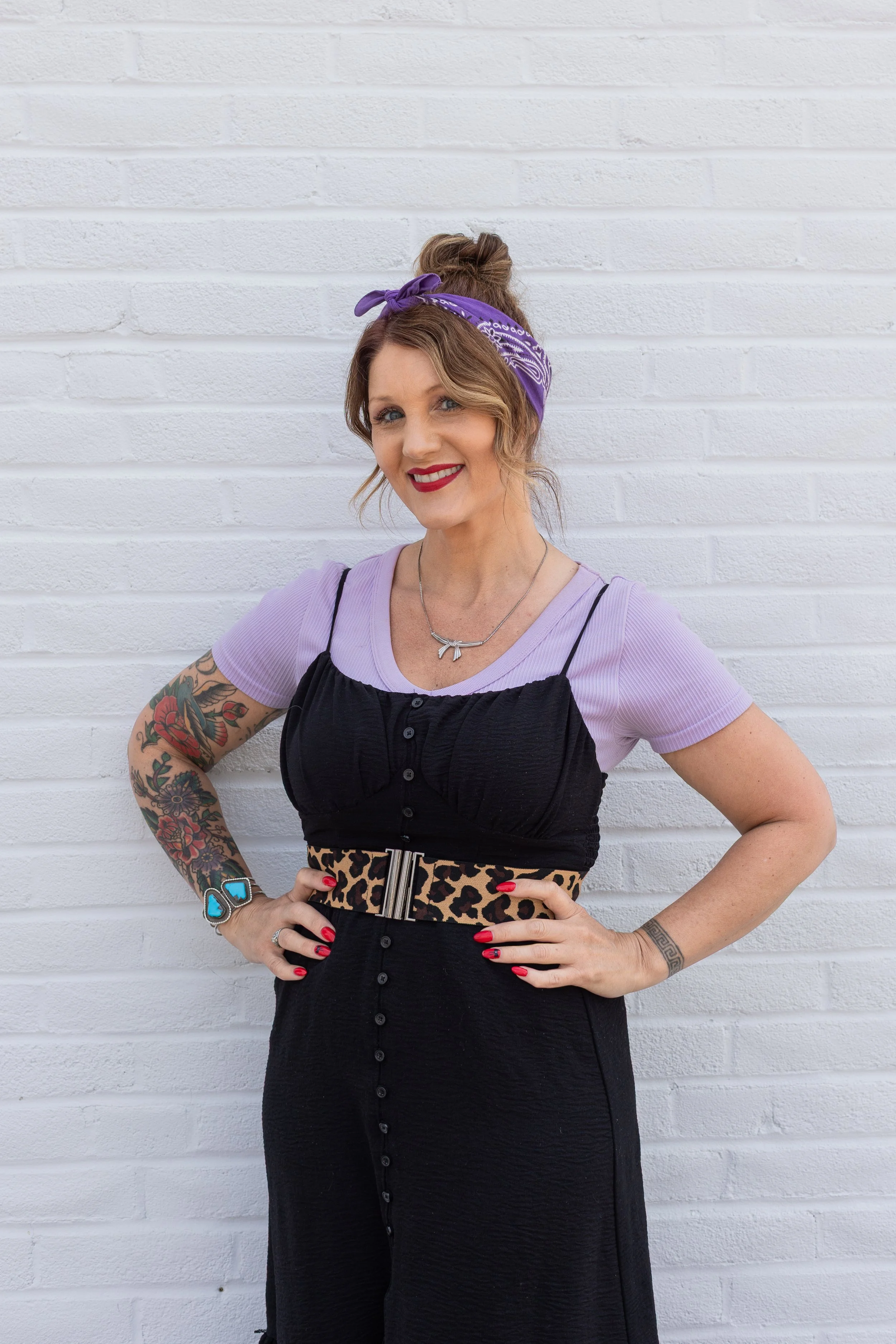 A woman with tattoos on her arms, wearing a purple bandana, a purple shirt under a black dress, a leopard print belt, and red nail polish, standing in front of a white brick wall, smiling at the camera.