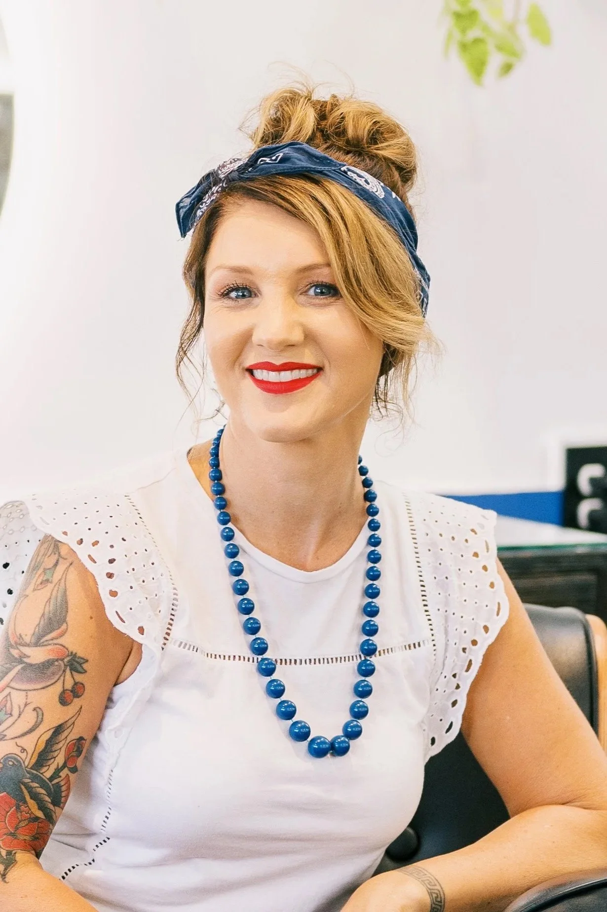 A woman with light skin and red hair, styled in vintage curls under a blue bandana, smiles while sitting at a hair station. She wears a white eyelet top, a blue bead necklace, and has tattoos on her left arm.