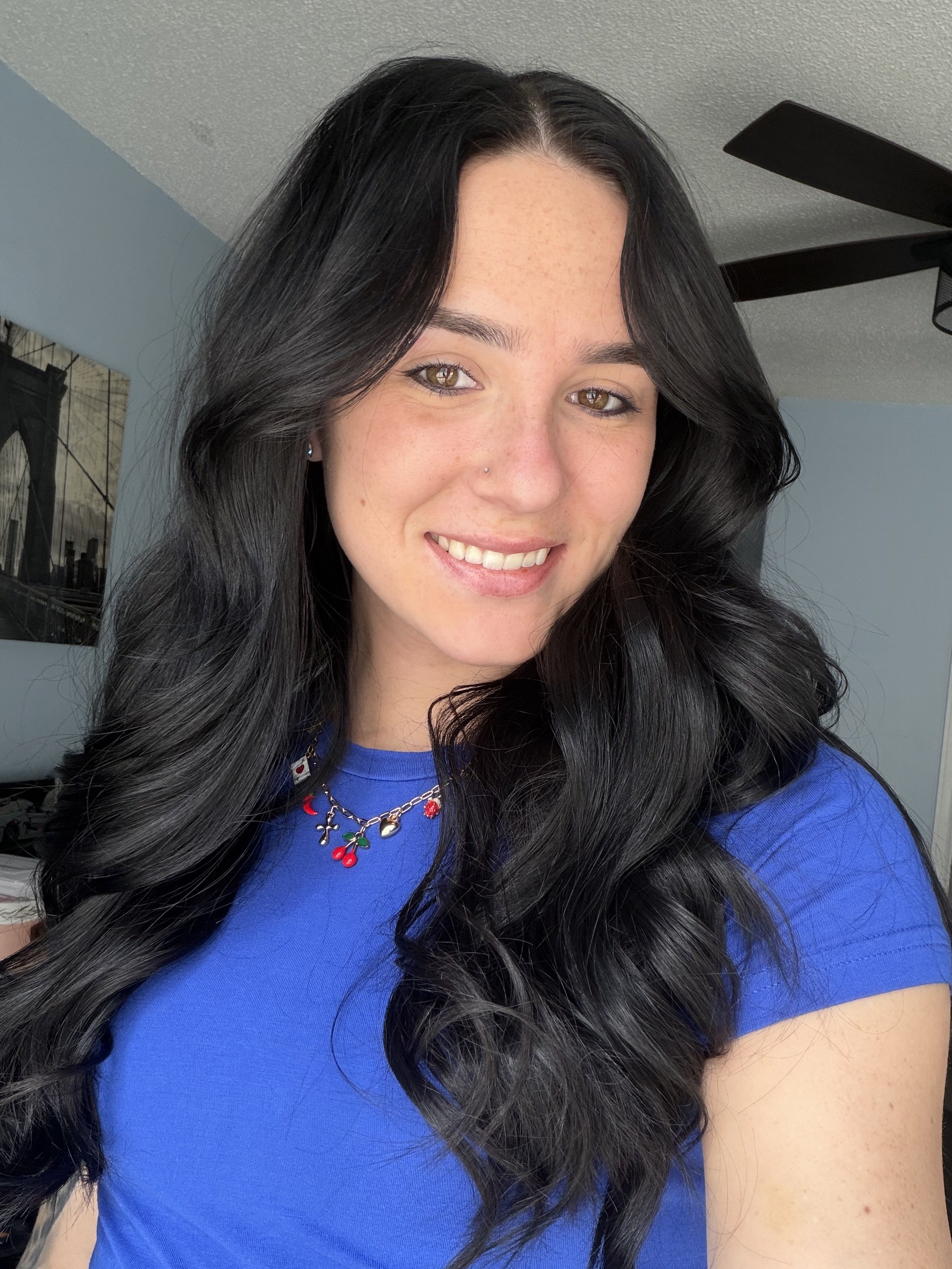 A woman with long, wavy black hair smiling at the camera. She is wearing a blue top and a necklace with cherries and crosses. She has brown eyes and a small nose piercing.