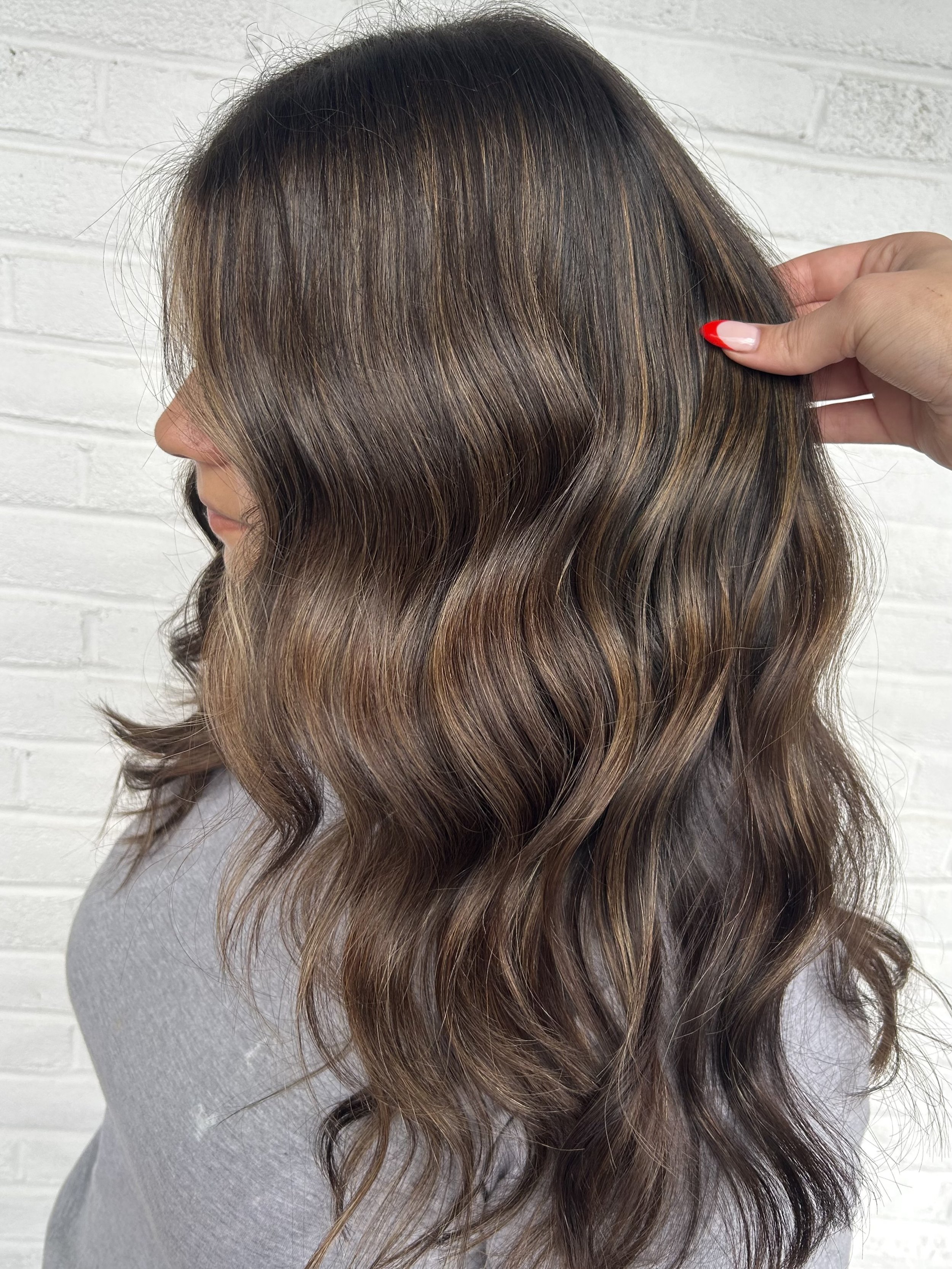 Side view of a woman with medium-length wavy hair in shades of brown with highlights, holding her hair back with her right hand against a white brick wall background.