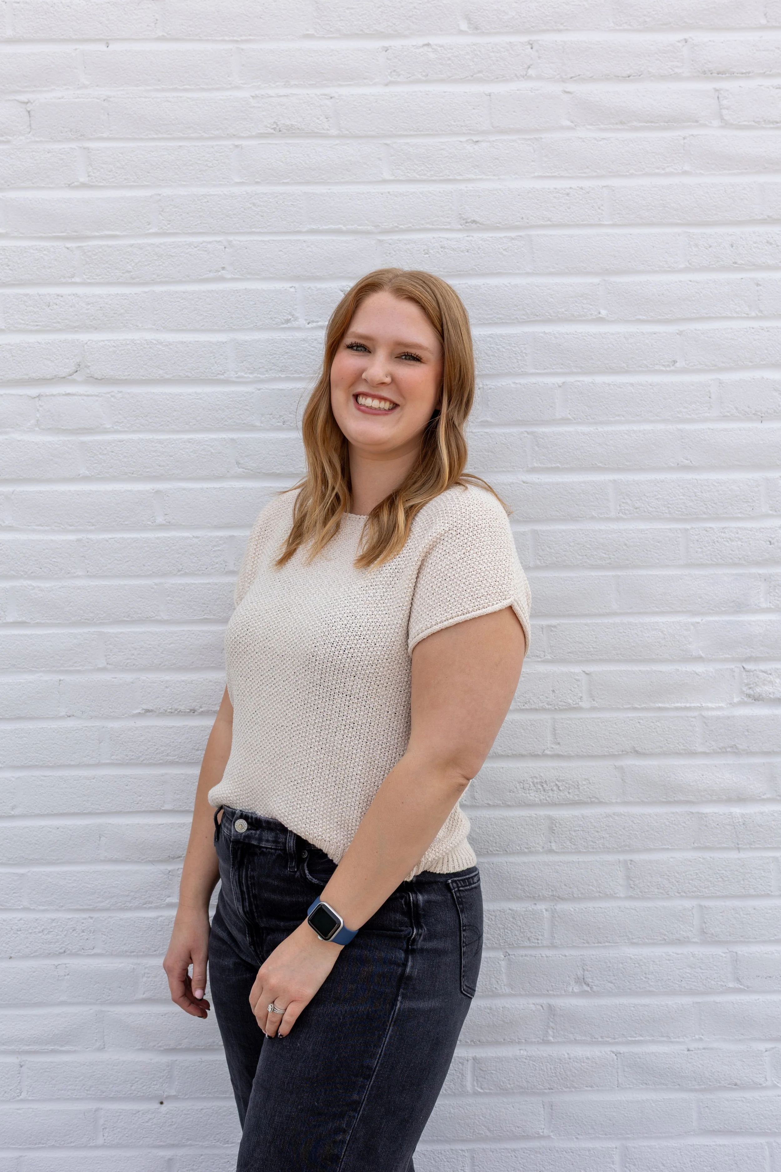 A woman with shoulder-length light brown hair, smiling, standing against a white brick wall, wearing a beige short-sleeved knit top, black jeans, and a smartwatch on her left wrist.