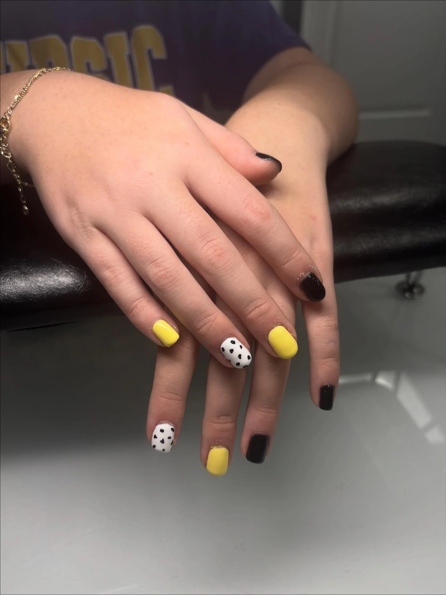 Close-up of a person's hand with black and yellow nail polish, resting on a gray surface, wearing a gold bracelet.