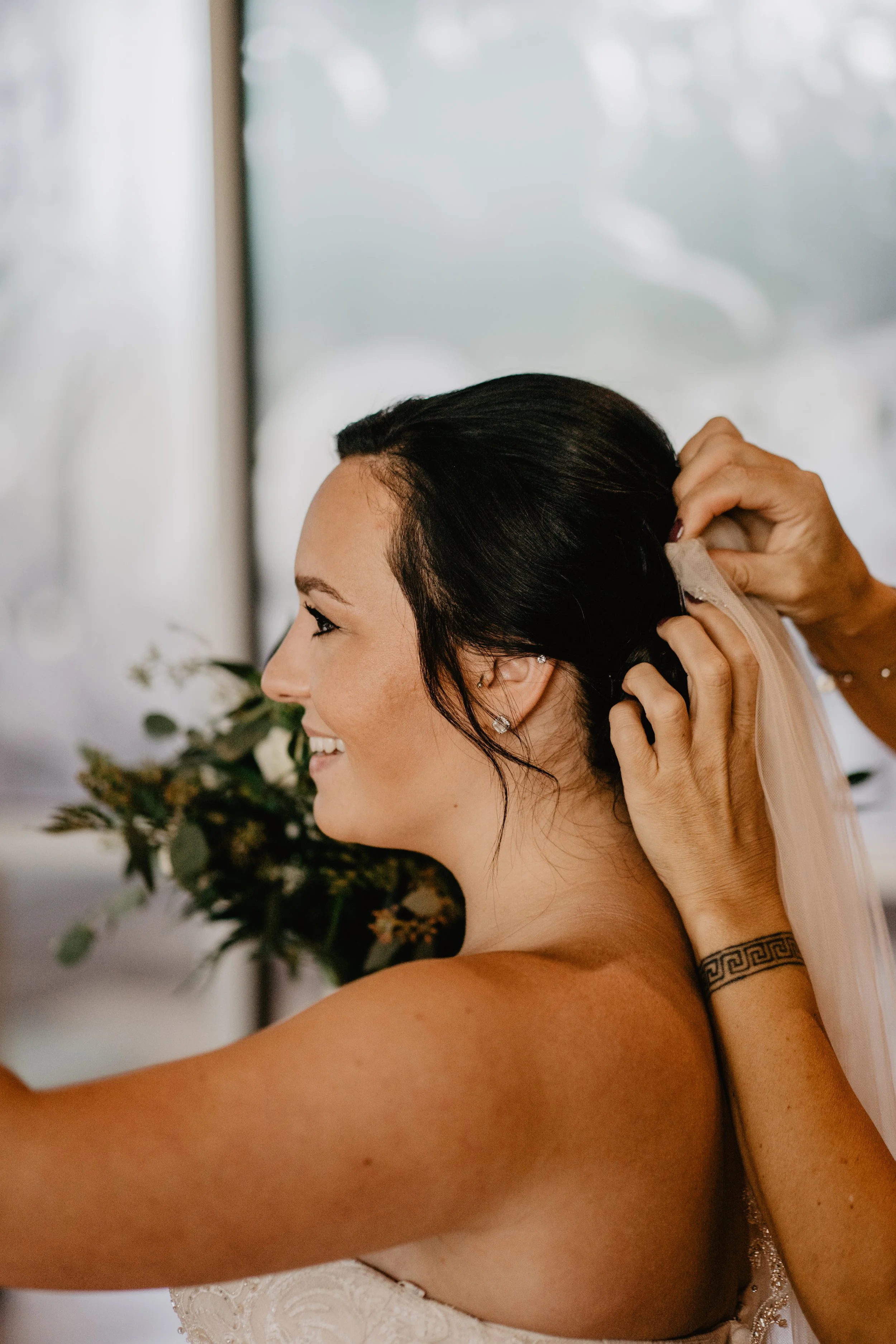 A woman with dark hair and earrings is smiling while someone adjusts her wedding dress near her shoulder on a wedding day.
