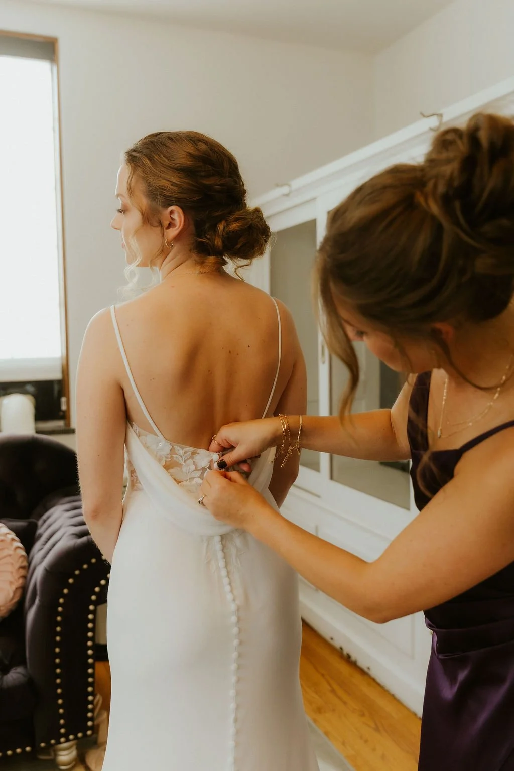 A woman in a white wedding gown with floral lace on the back is being assisted by another woman with fastening buttons on the back of her dress.