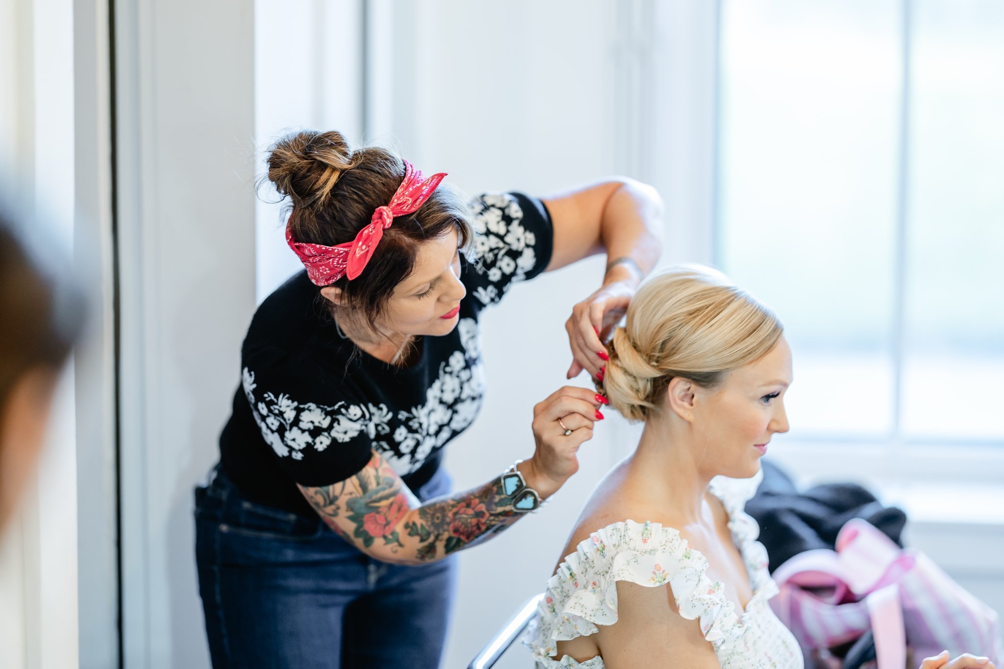 A hairstylist with tattoos and a red bandana hair accessory styles a woman's blonde hair in a braid in a bright room with large windows.