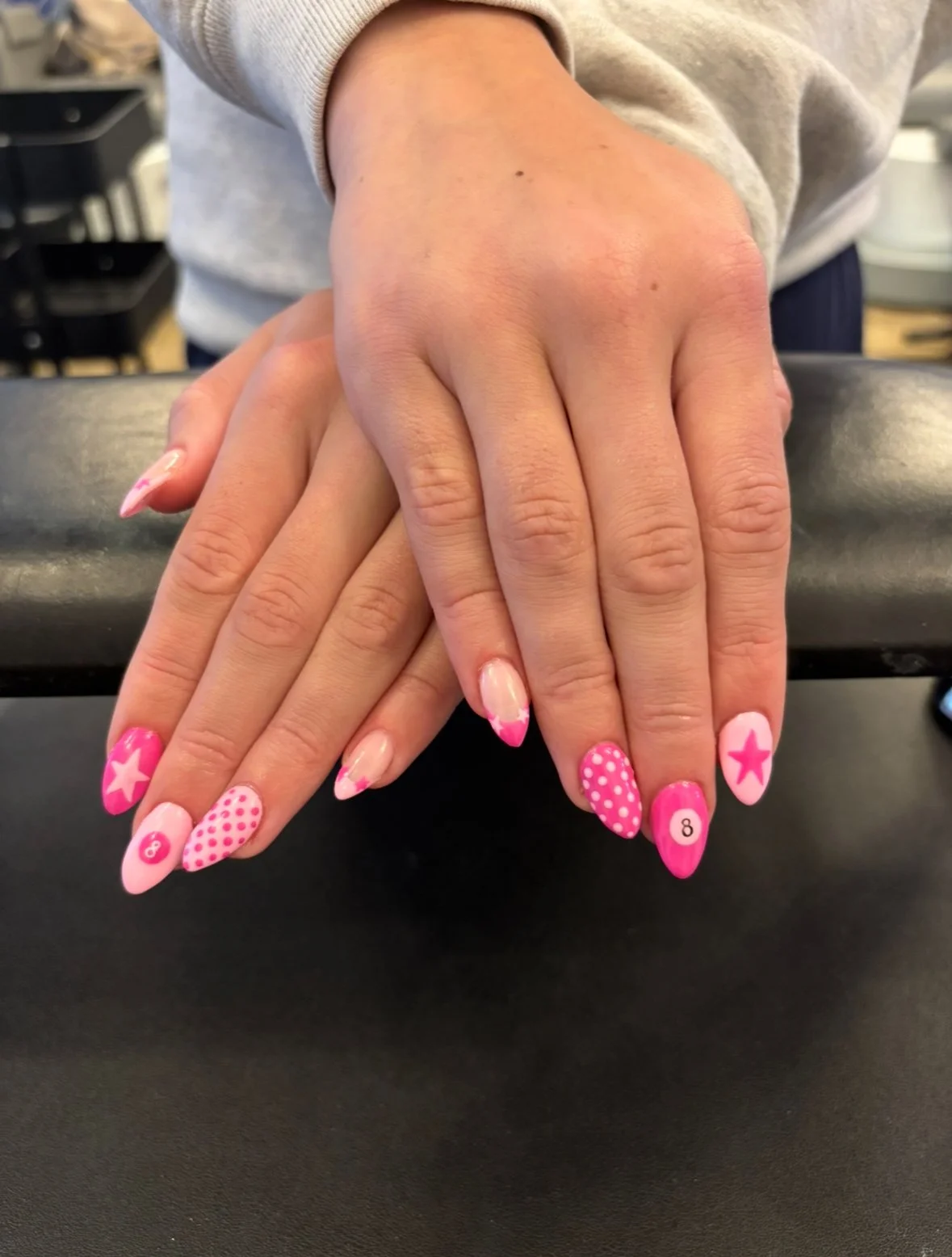 Close-up of two hands with pink and white decorated nails resting on a dark surface, with a blurred background.