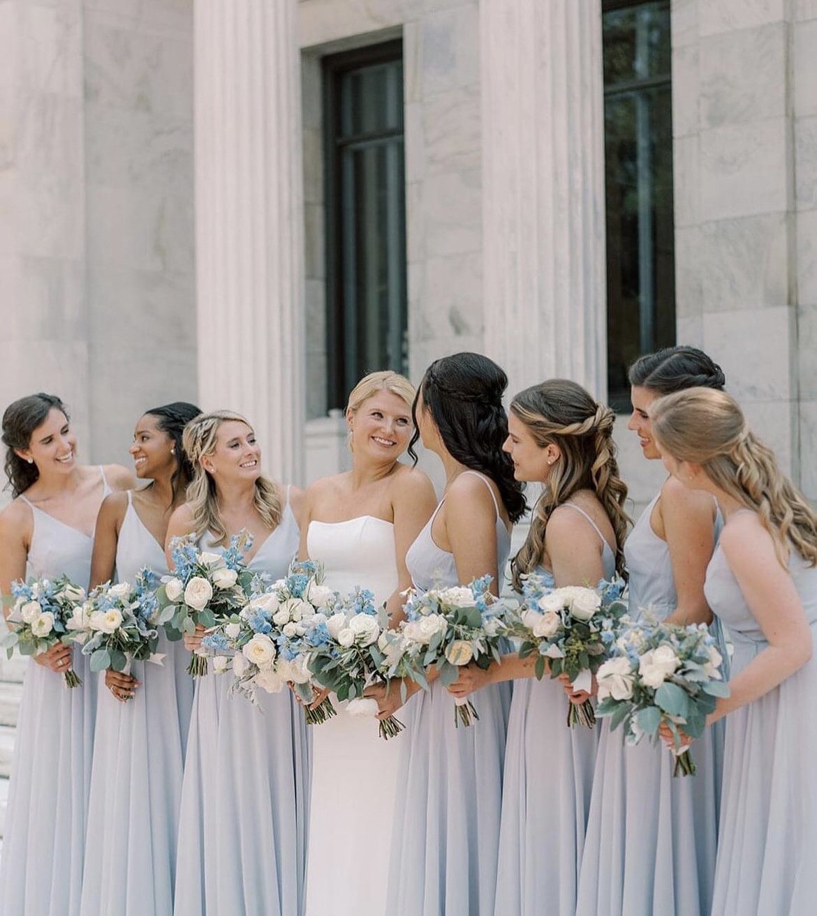 A group of women in bridesmaid dresses holding bouquets, gathered in front of a building with columns, celebrating at a wedding.