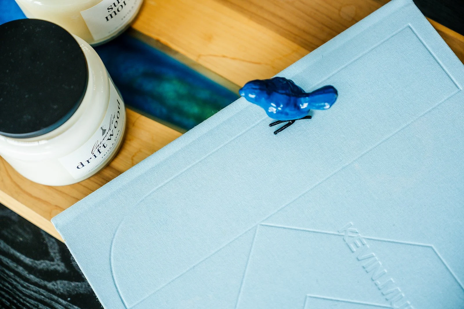 A blue decorative bird figurine perched on the corner of a light blue book, with a white candle jar labeled 'driftWOOD' on a wooden surface nearby.