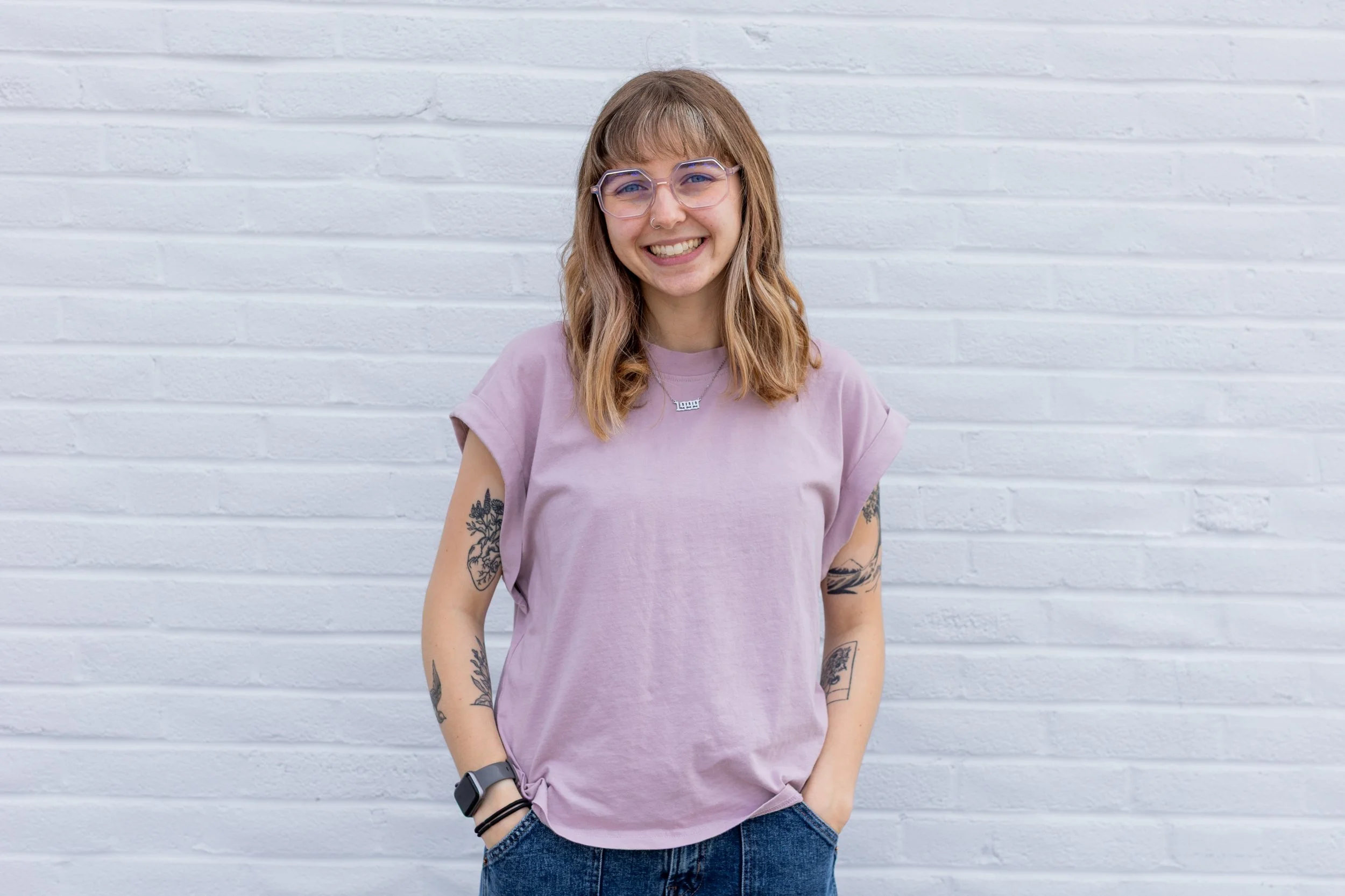 A woman with shoulder-length light brown hair and glasses, wearing a lavender T-shirt with rolled sleeves and blue jeans, standing in front of a white brick wall. She is smiling and has tattoos on her arms.